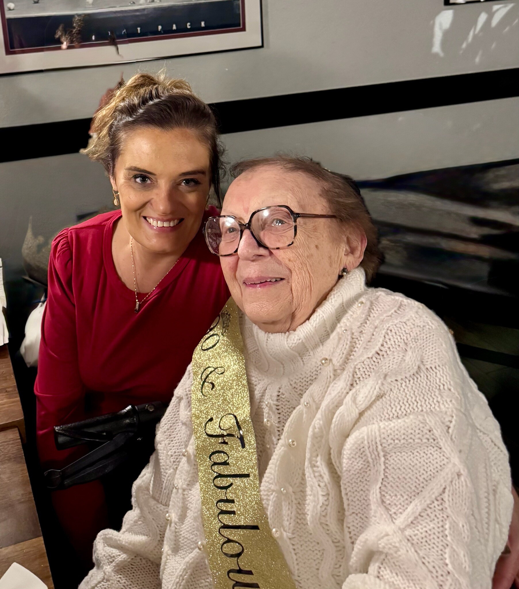 Two women smiling for a selfie, one in a red top and the other in a cream sweater with a gold glitter ribbon saying 'Congratulations' over her chest.