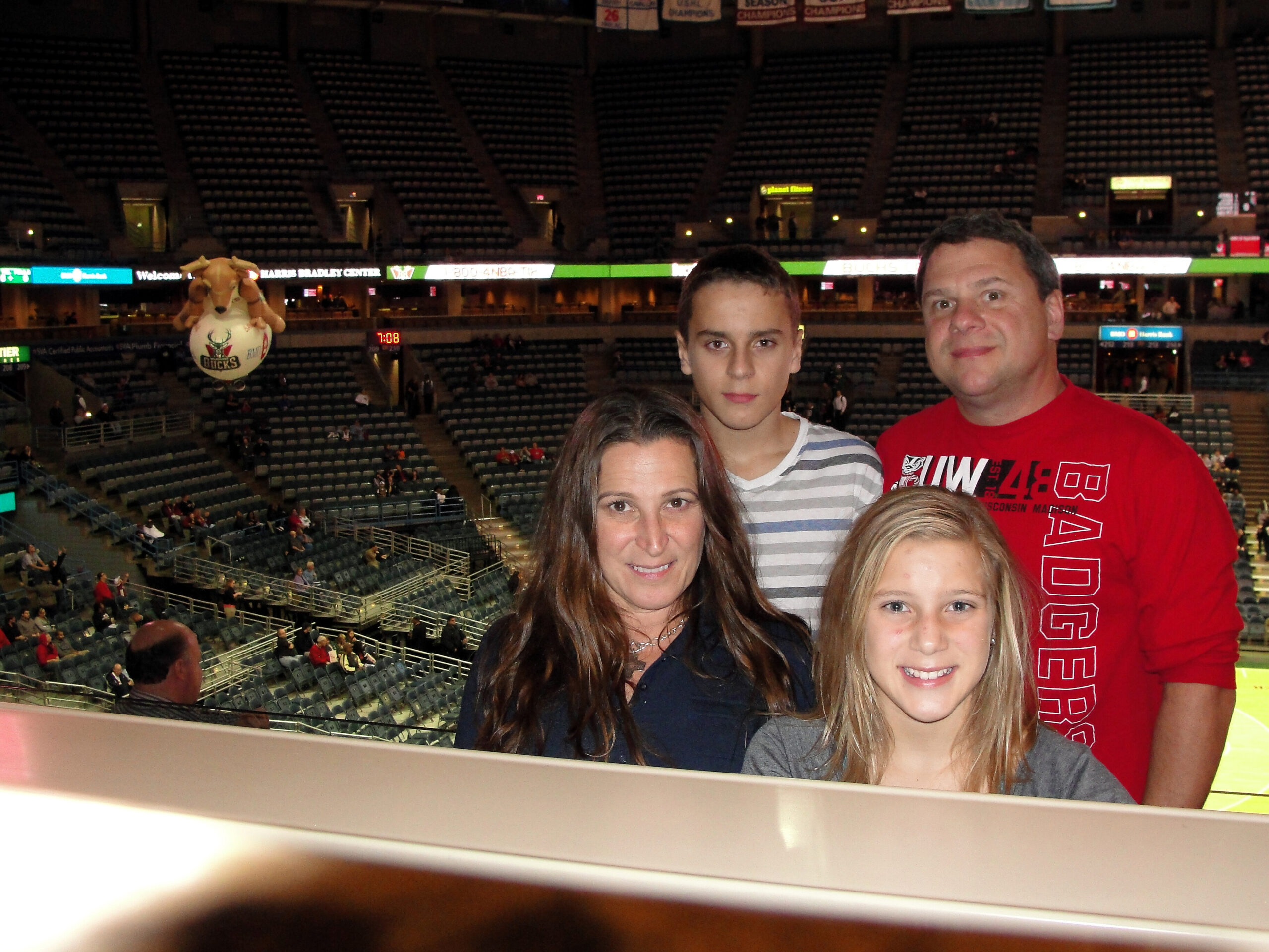 Family of four posing for a photo inside a large sports arena with empty stands and a Bucks mascot balloon in the background.
