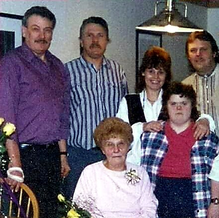 Multi-generation family posing indoors around a dining table; elderly woman in pink sits front center, others stand behind her.