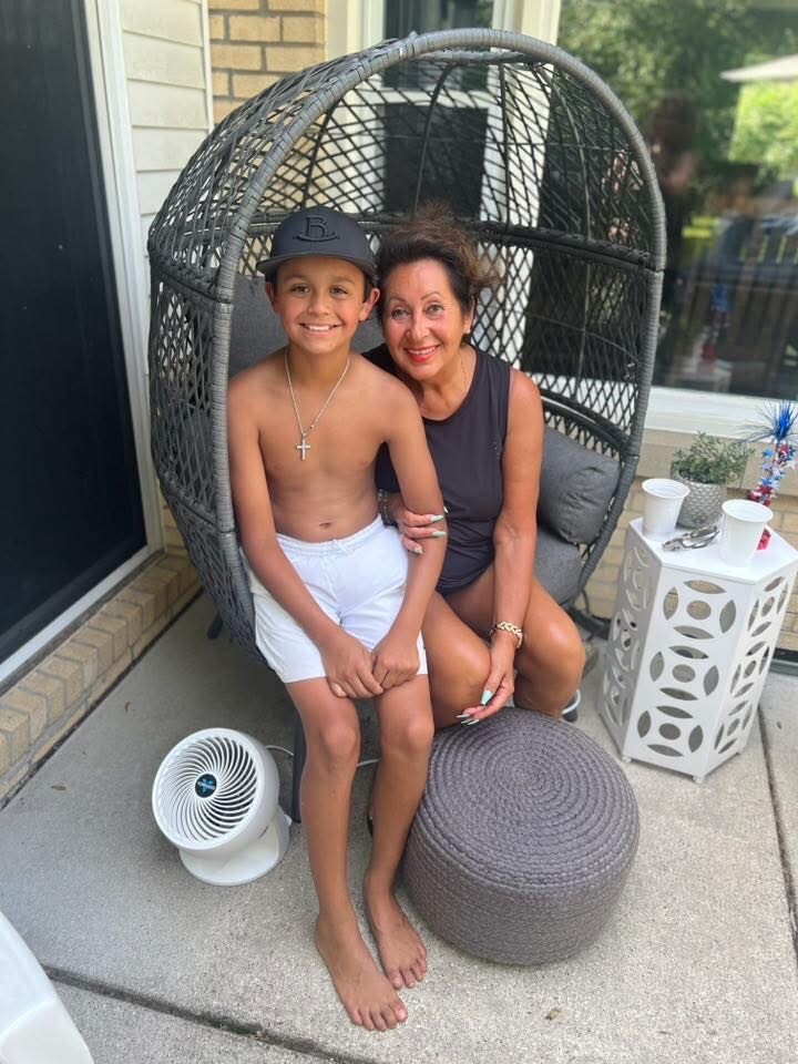 Smiling boy in white shorts and cap sits with a woman in a dark sleeveless top on a large wicker outdoor chair.
