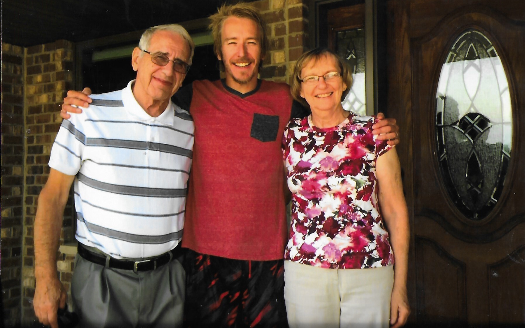 Three smiling people stand side by side on a porch, with a brick wall and wooden door behind them (two older adults flanking a younger man).
