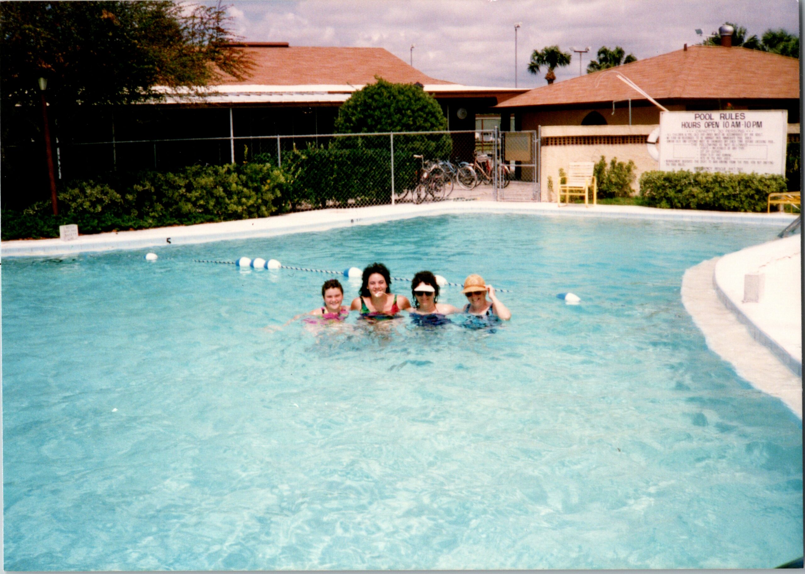 Four people smiling in a sparkling blue pool near the edge, with a chain-link fence and a pool rules sign in the background.