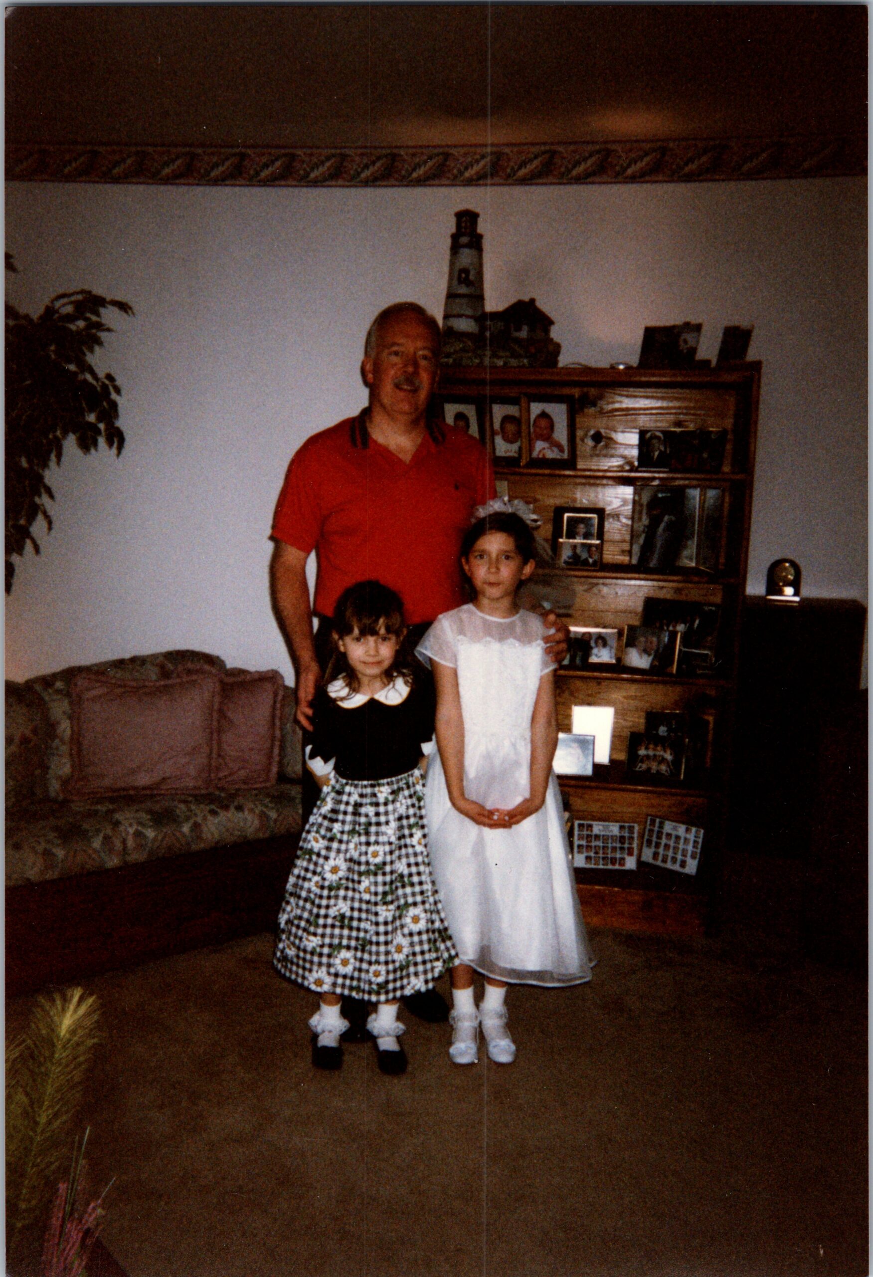 Adult man in a red polo stands behind two young girls in dresses in a living room, posing for a family photo.