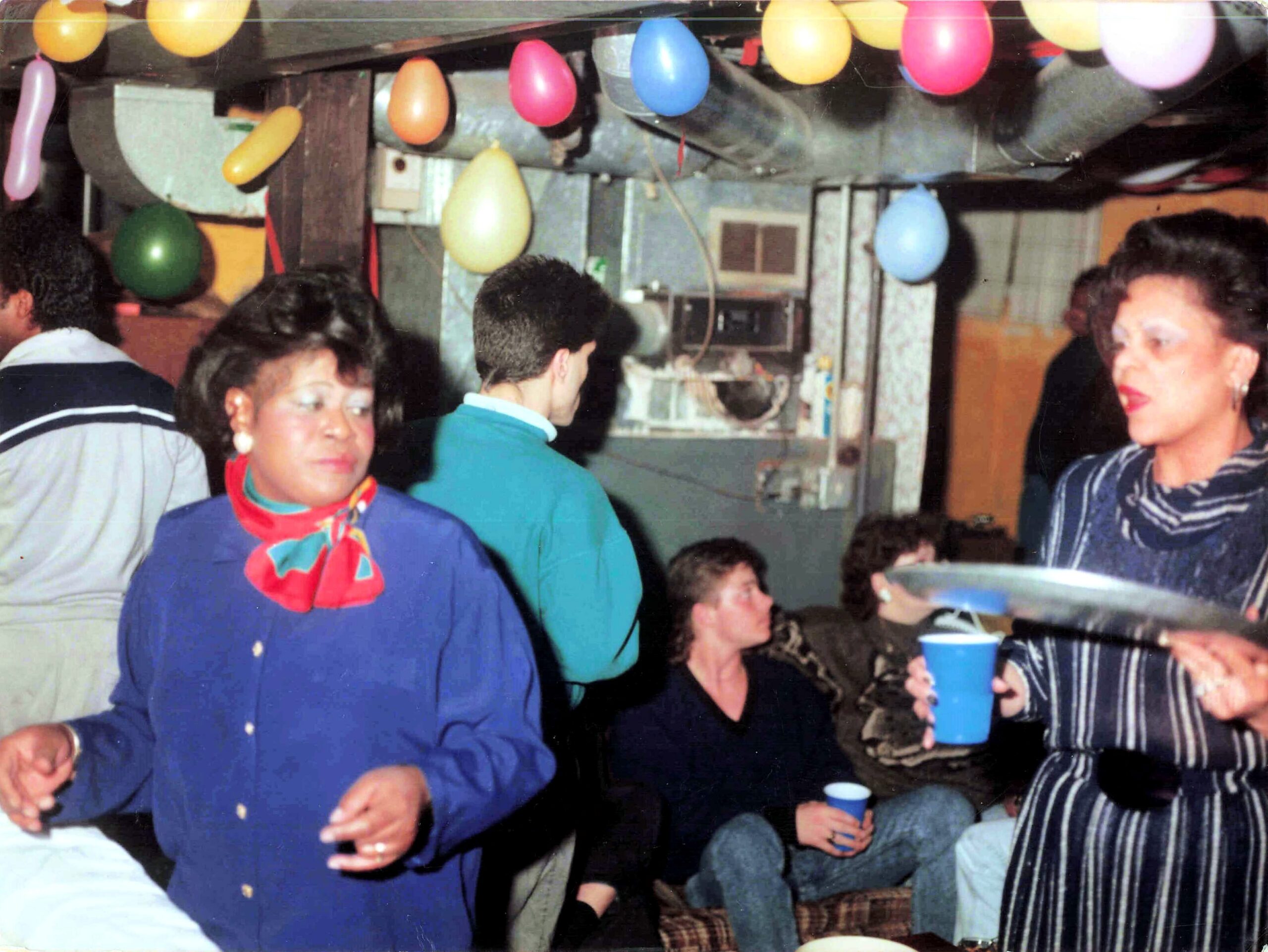 People at a festive party indoors with colorful balloons overhead; a woman in a blue outfit with a red scarf stands in the foreground while others mingle and hold blue cups.