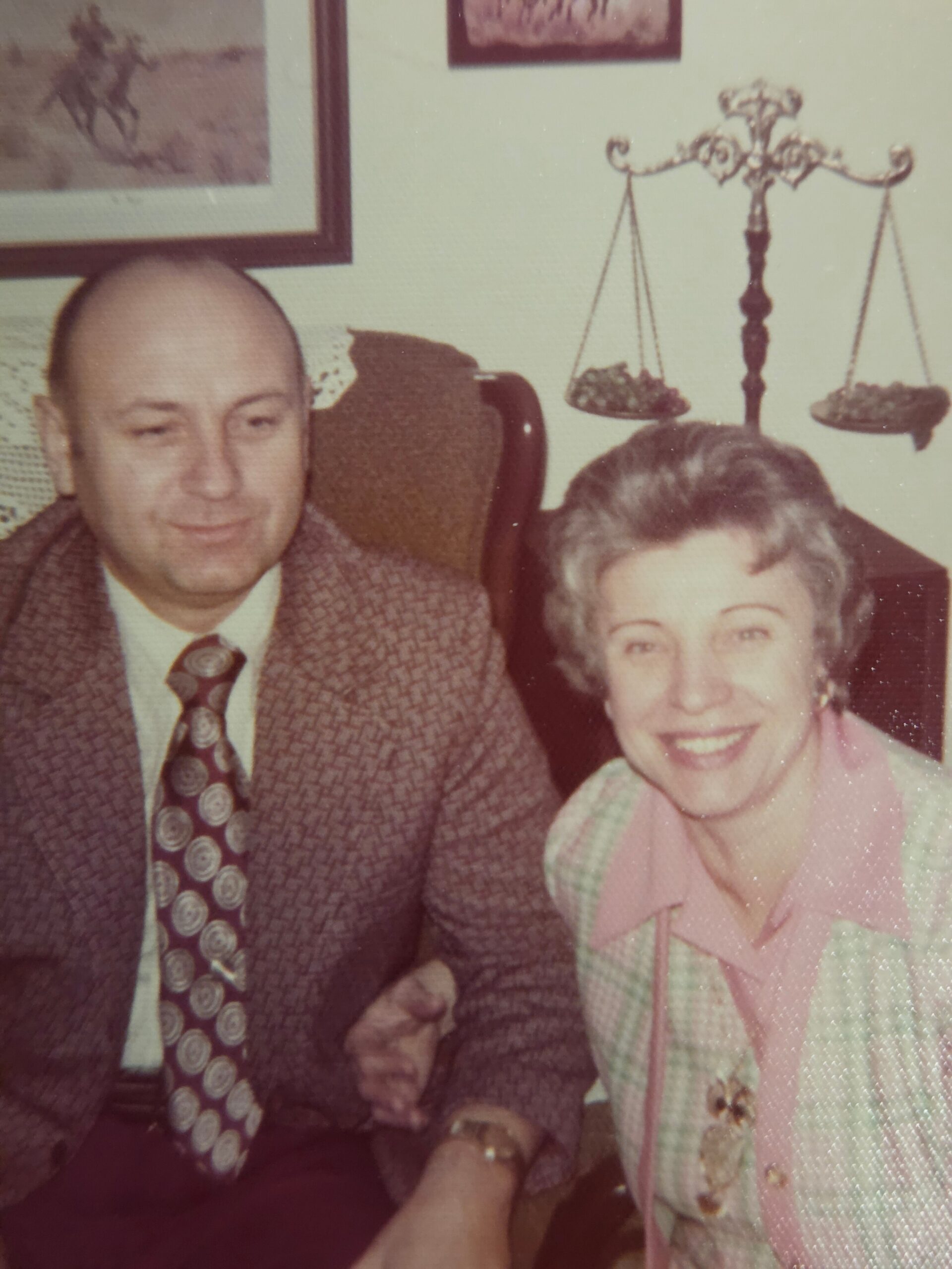Smiling elderly couple seated together; man in a brown patterned suit and tie, woman in a pink jacket, with an ornate scale decoration in the background.