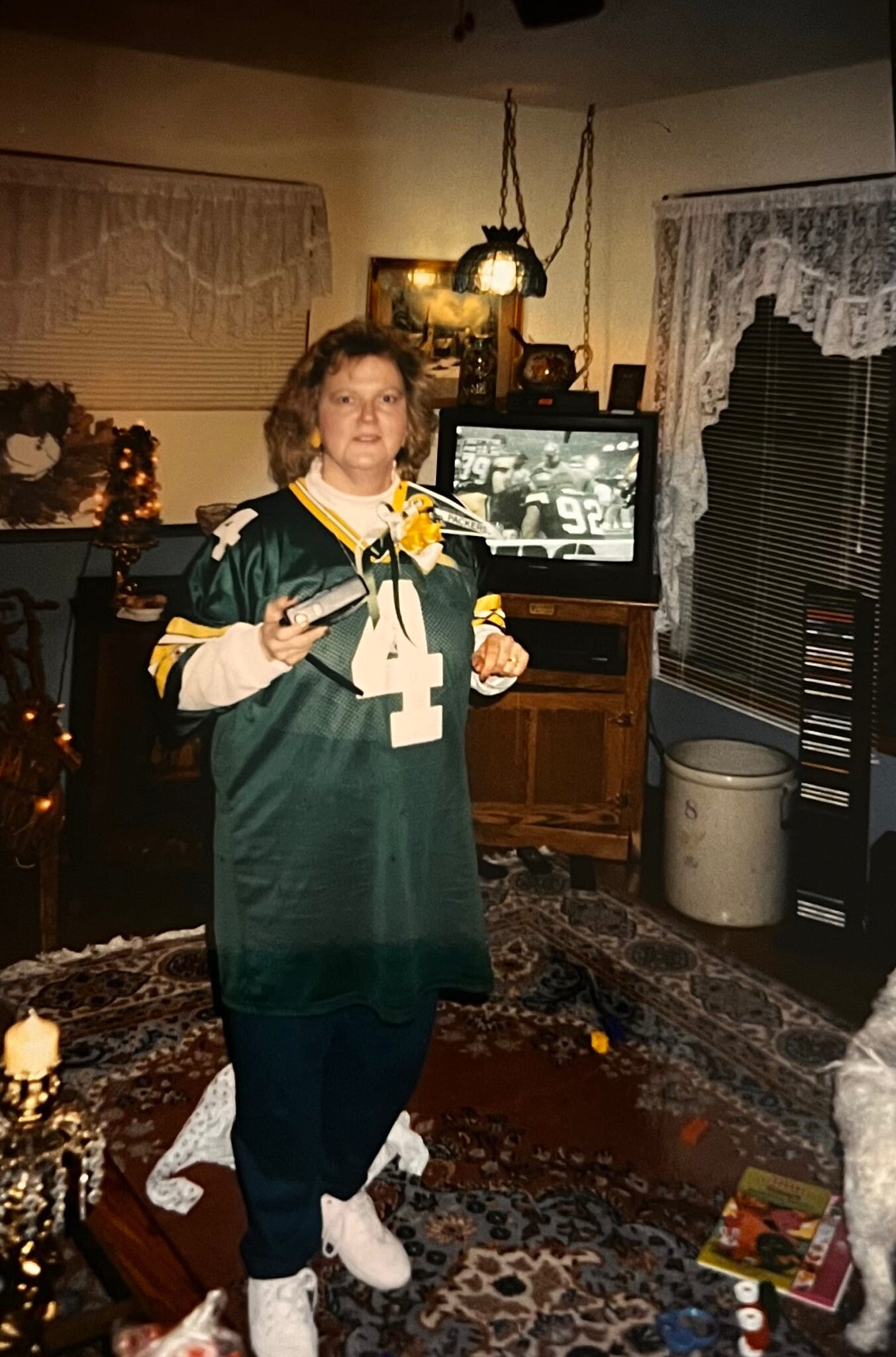Woman in a green sports jersey with the number 4 stands in a living room, holding a remote with a TV showing a football game behind her.