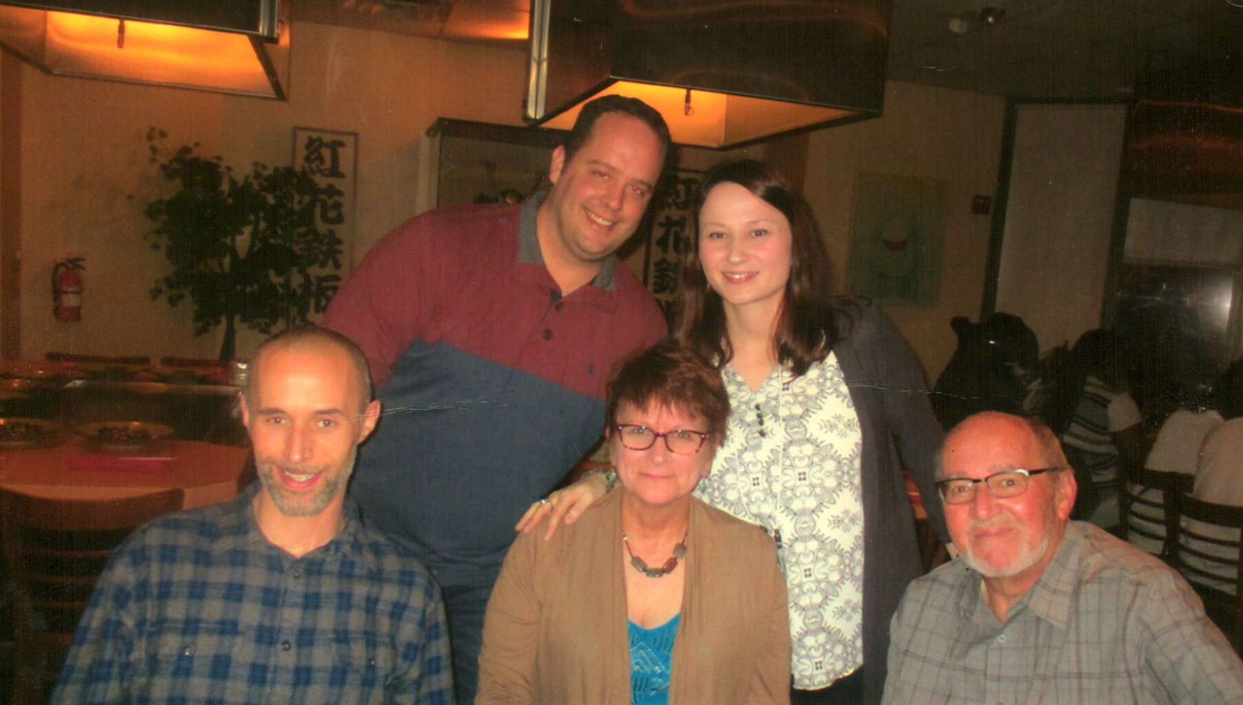 Group of five adults smiling for a photo in a restaurant, with two men seated in front, a woman in the middle, and two people standing behind them in casual attire.