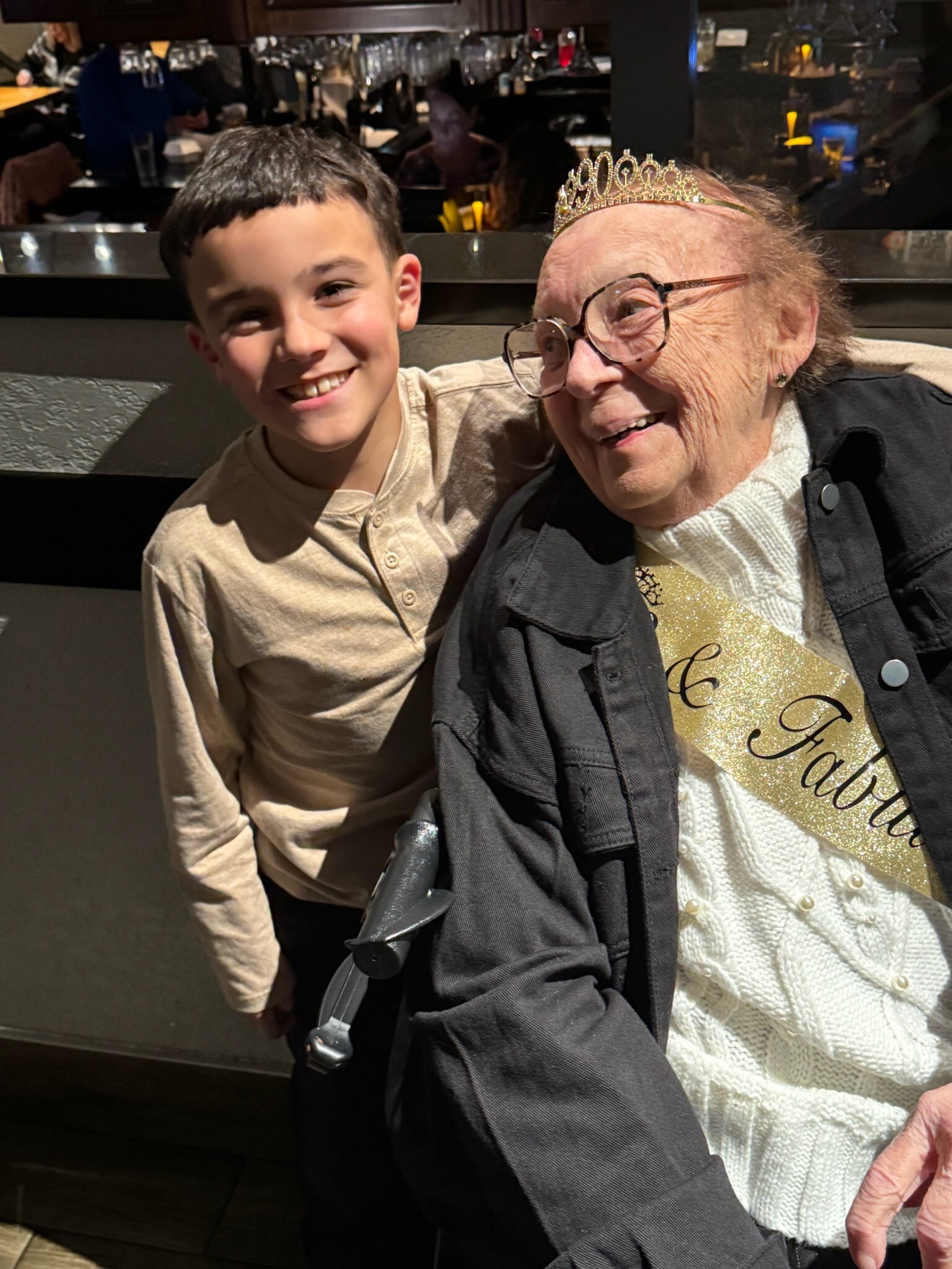 Smiling boy beside an elderly woman wearing a gold crown and glittery sash at a restaurant.