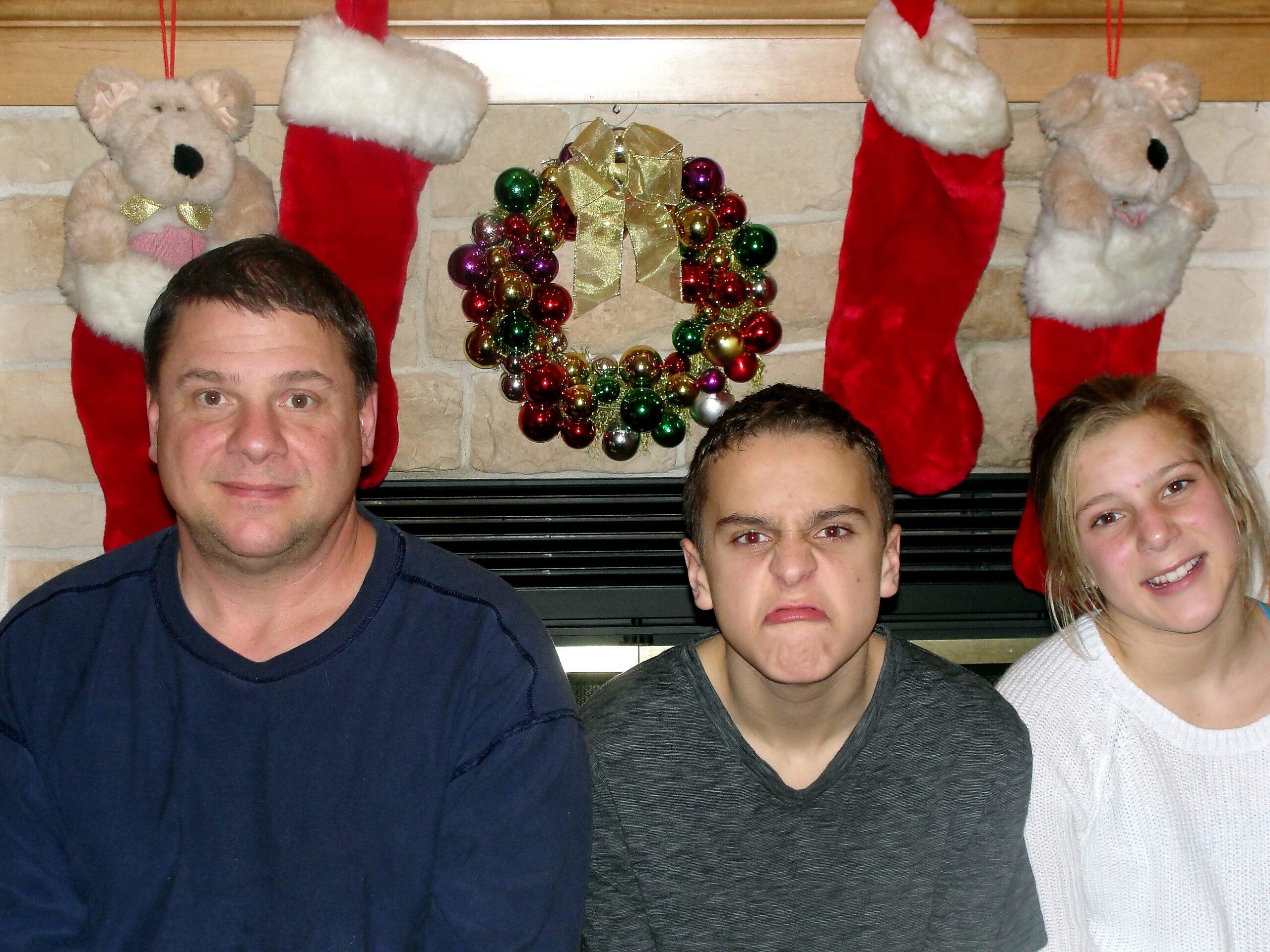 Family posing for a Christmas photo in front of a mantle with stockings, a wreath, and festive decor in the background.