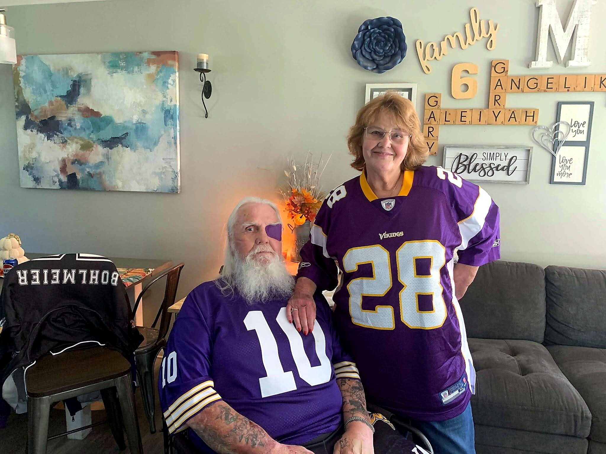 Smiling woman in a purple Minnesota Vikings jersey stands next to an older man with a long white beard in a living room with blue abstract art and wall décor behind them.