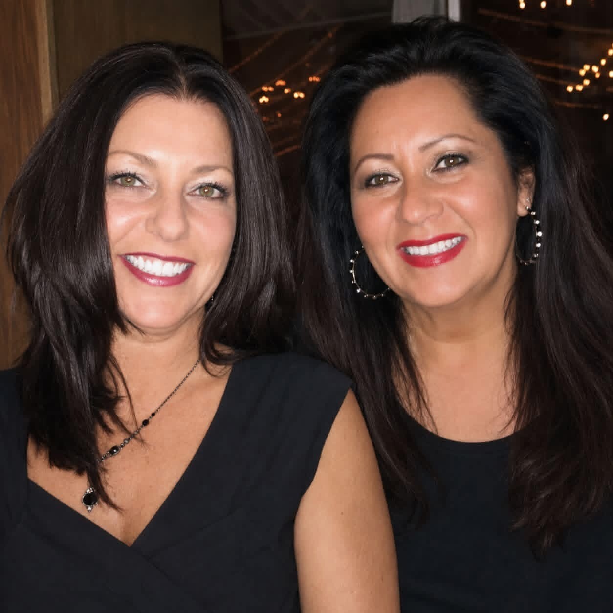 Two women with dark hair smiling at the camera, wearing black dresses, in a warmly lit indoor setting with string lights in the background.