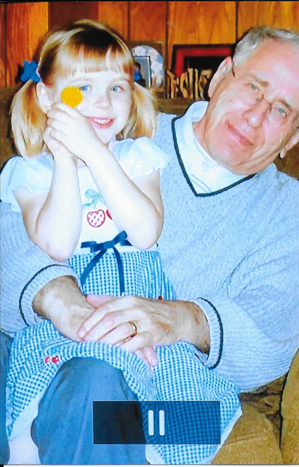 Grandfather and granddaughter posing on a couch; girl playfully covers one eye with a yellow pom-pom and smiles.
