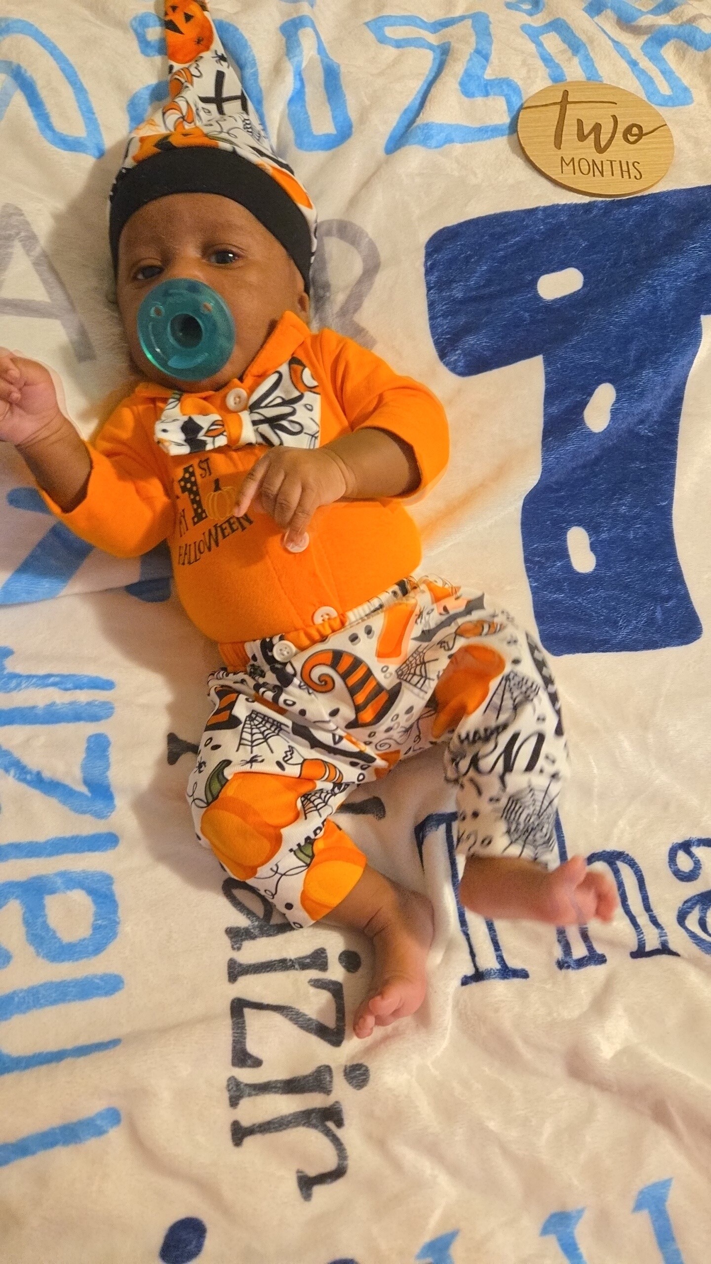 Infant in orange Halloween outfit with bow tie and teal pacifier, lying on a white blanket with blue patterns; a small round 'Two months' badge nearby.