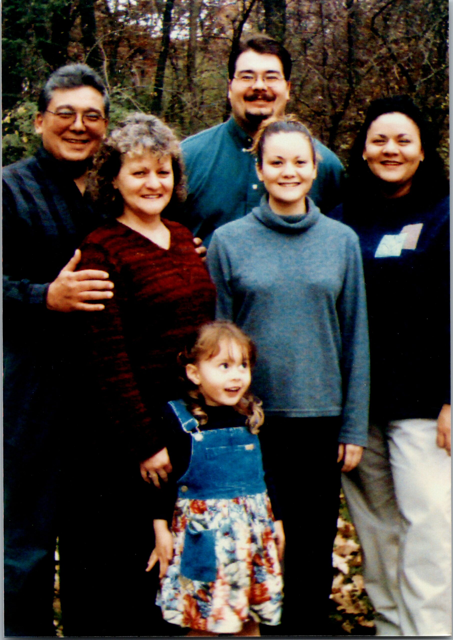 A group of five people smiling outdoors in a fall wooded setting, with a young girl in a blue dress in front.