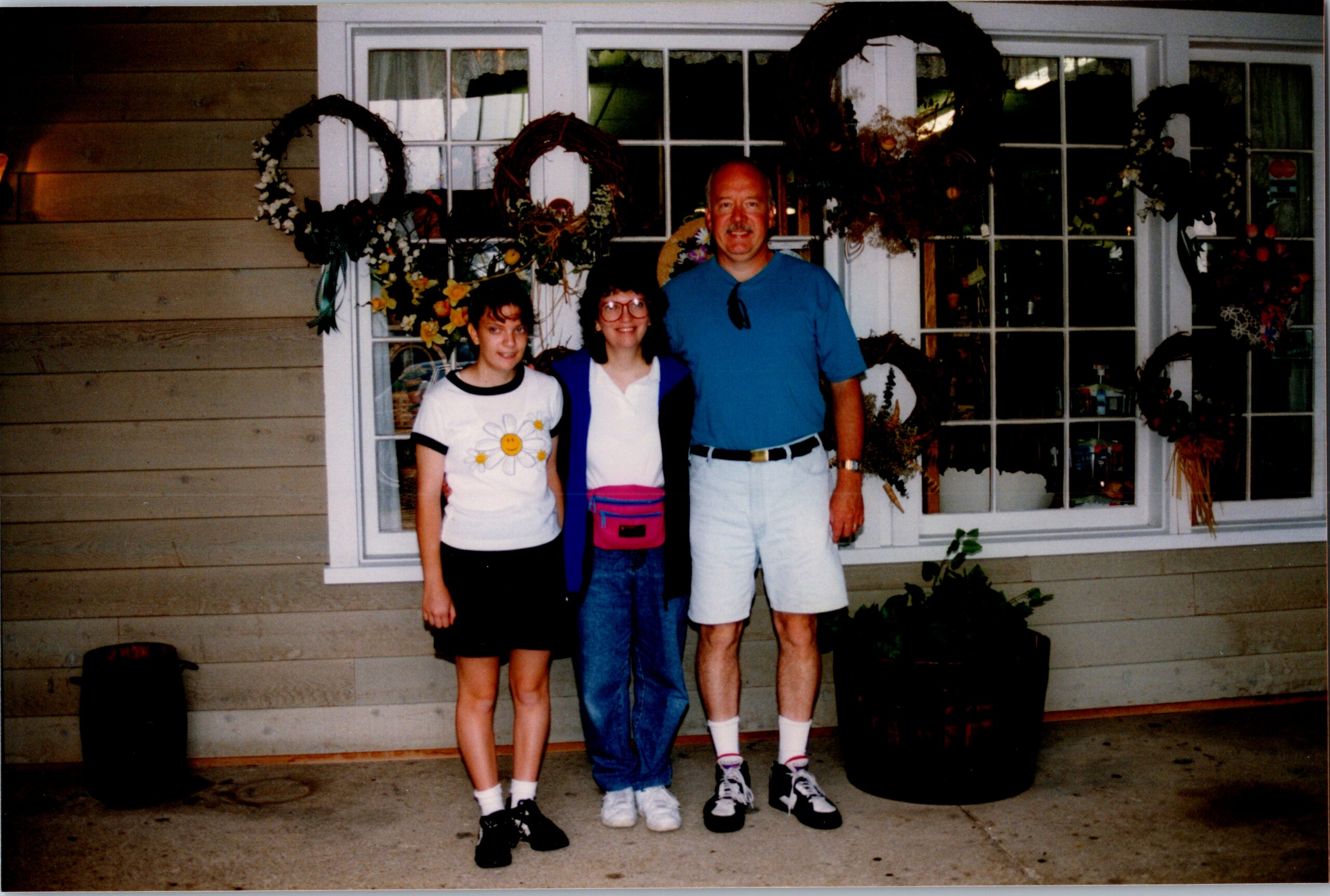 Three people posing together outside a house in front of a window decorated with wreaths and flowers.