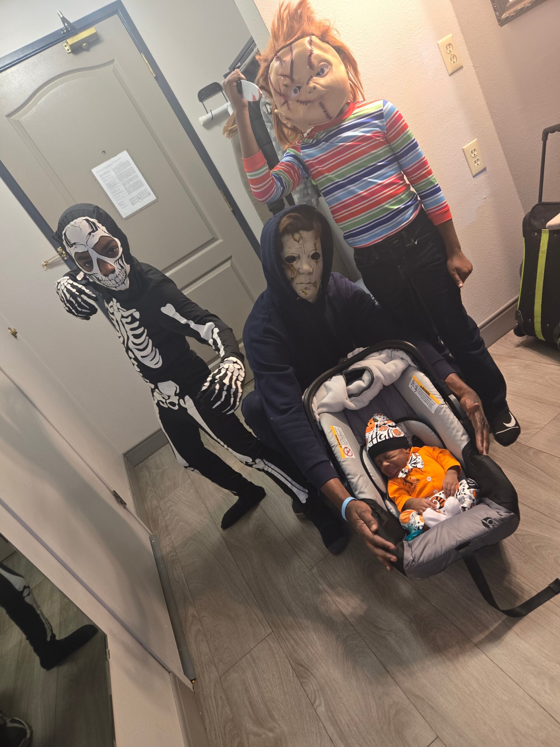 Kids in Halloween costumes with eerie masks pose in a hallway, one in a striped shirt and a stitched face mask, another in a skeleton suit, standing beside a baby in a car seat in a stroller area