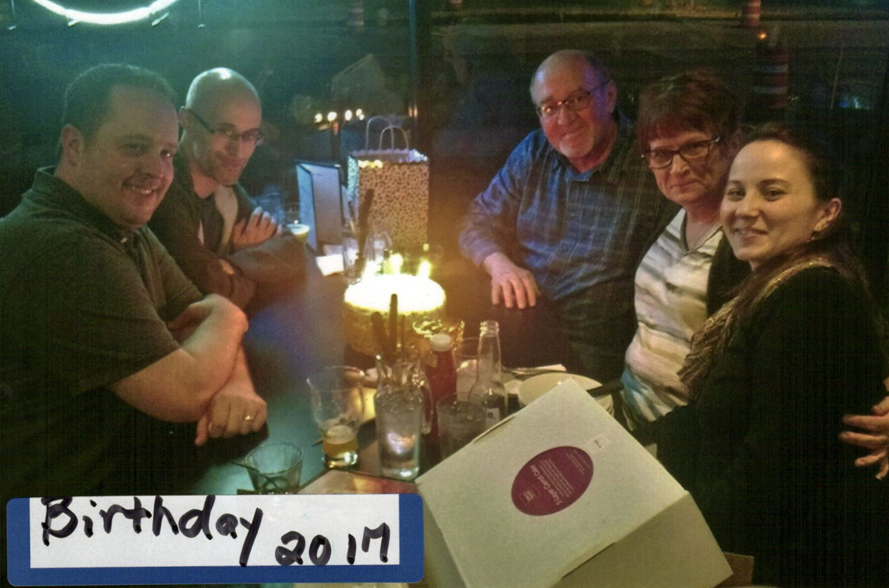 Five adults sit around a candlelit restaurant table for a birthday celebration, with a glowing cake in the center and drinks and gifts on the table.