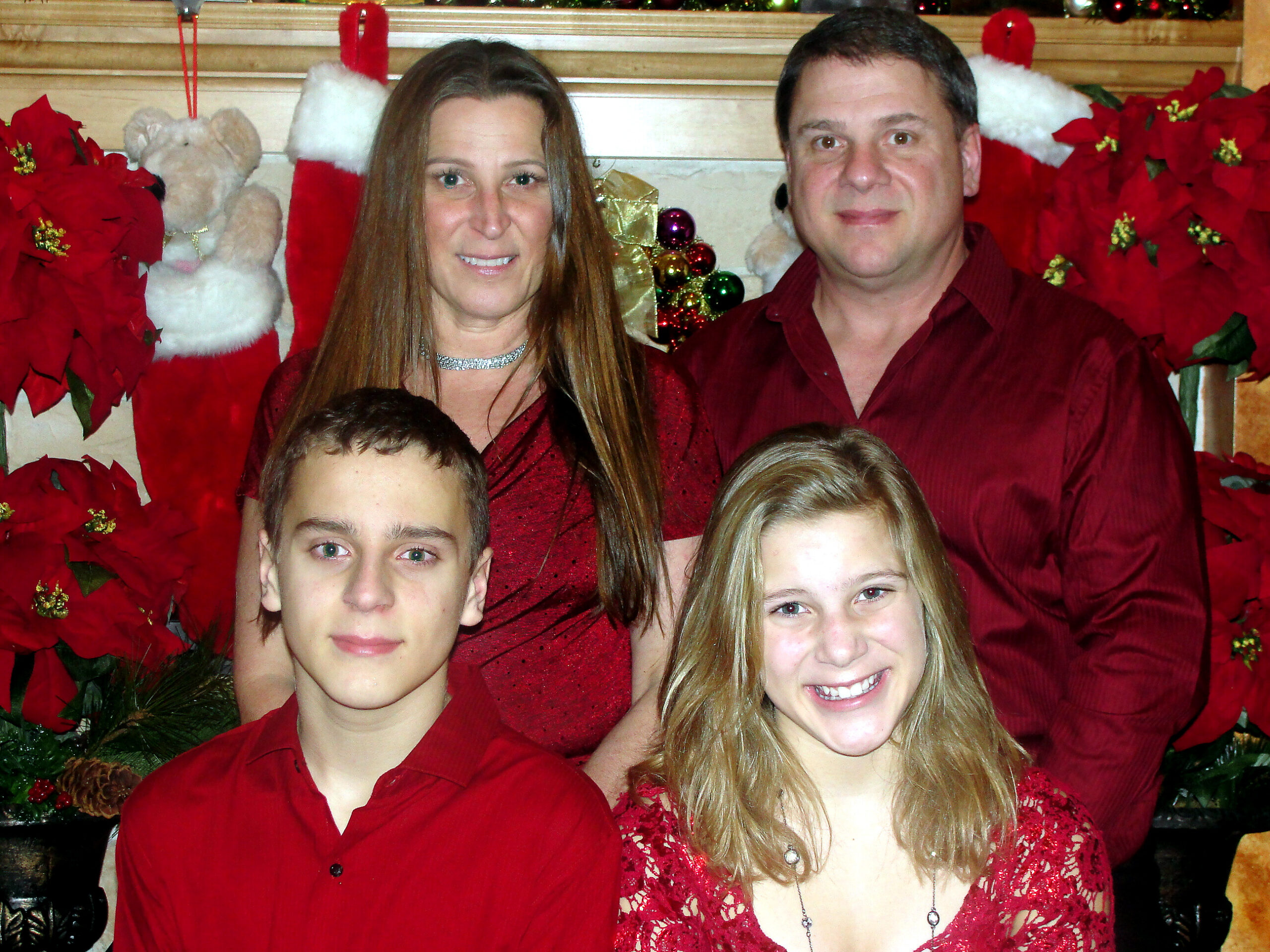 Family posing for a Christmas photo in red outfits with poinsettias and ornaments in the background, smiling at the camera.