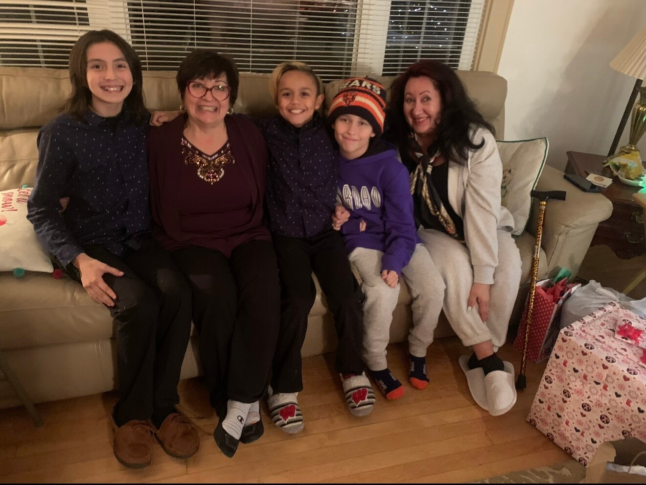 Five people sit closely on a sofa, smiling for the camera, including two adults and three children, in a cozy living room with gift bags nearby.