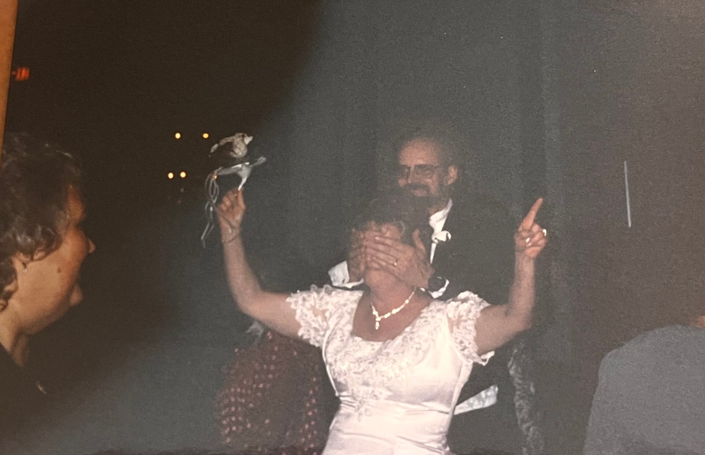 Woman in a white dress dancing with arms raised at a night-time party, warm lights in the background with other guests nearby.