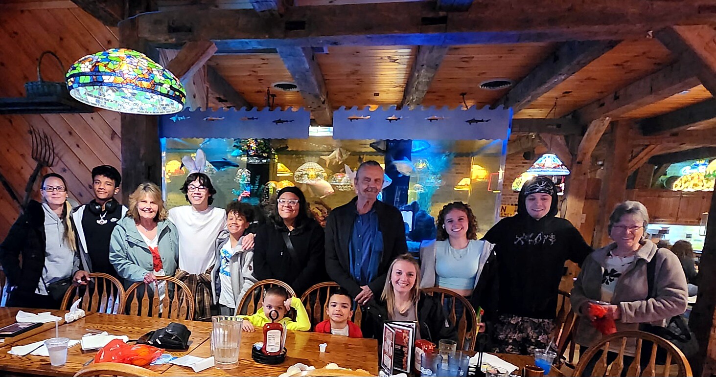Group of people of various ages posing for a photo around a long wooden dining table inside a rustic restaurant, with an aquarium display in the background.