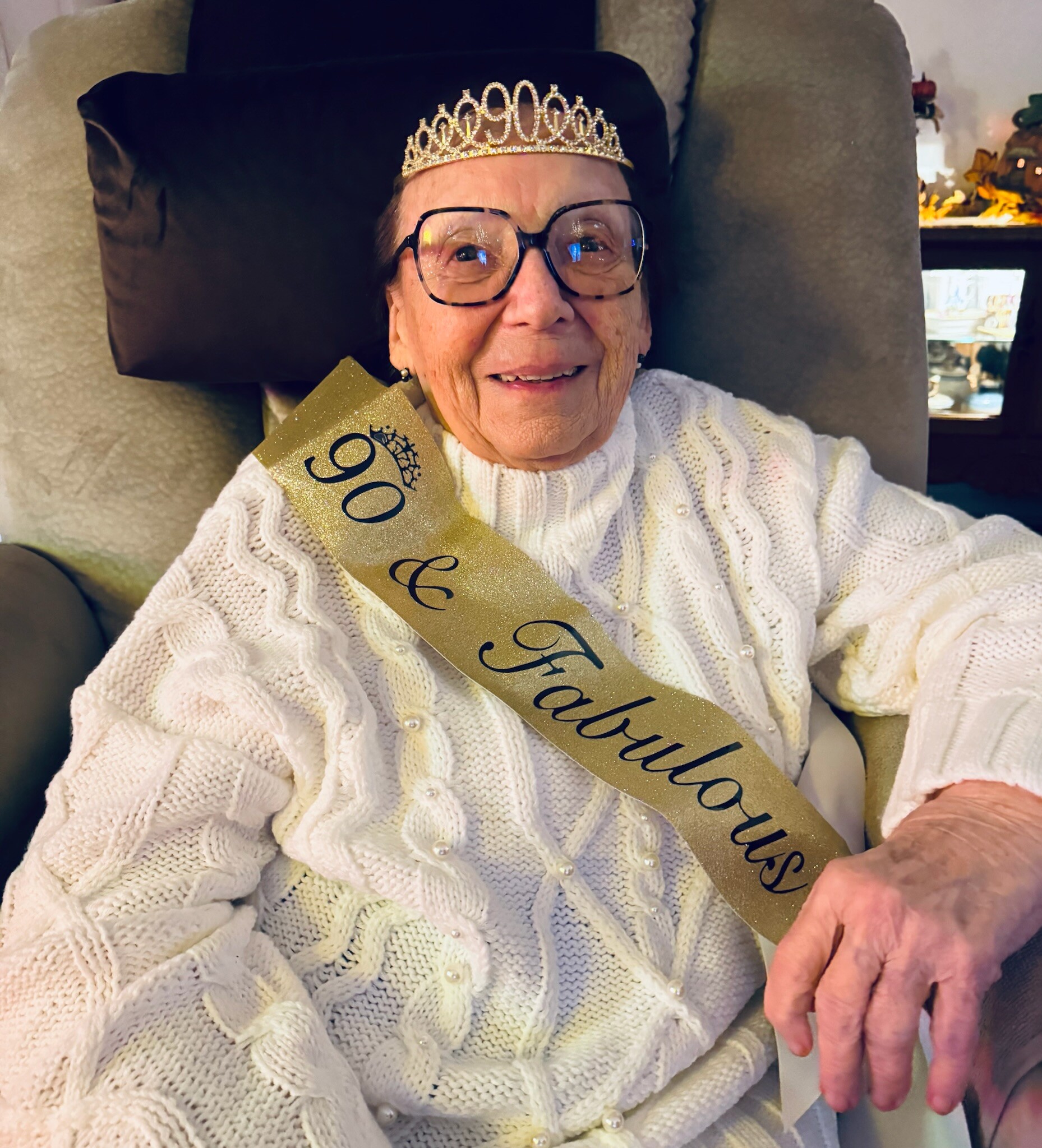 Elderly woman wearing a tiara and glasses, smiling while seated in a chair with a gold birthday sash across her chest.