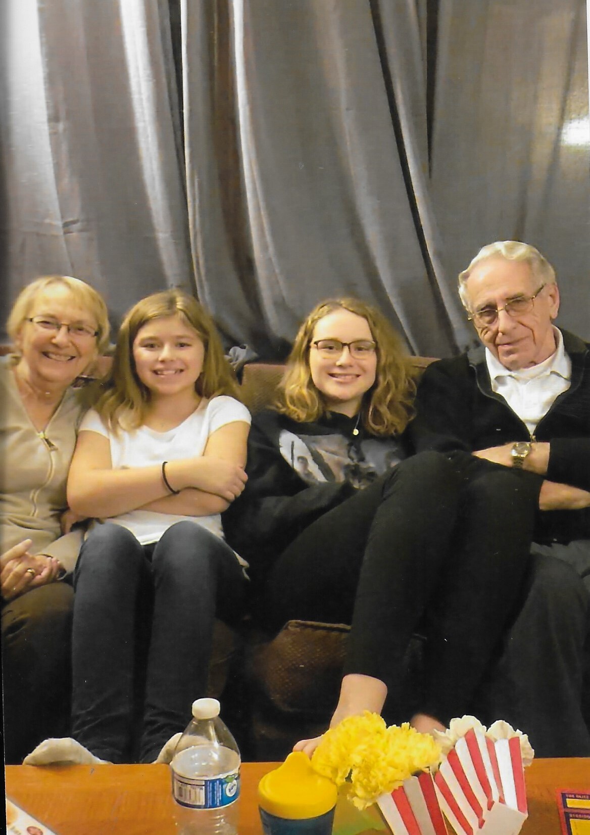 Four people—a woman, two girls, and an elderly man—sit together on a couch, smiling at the camera.