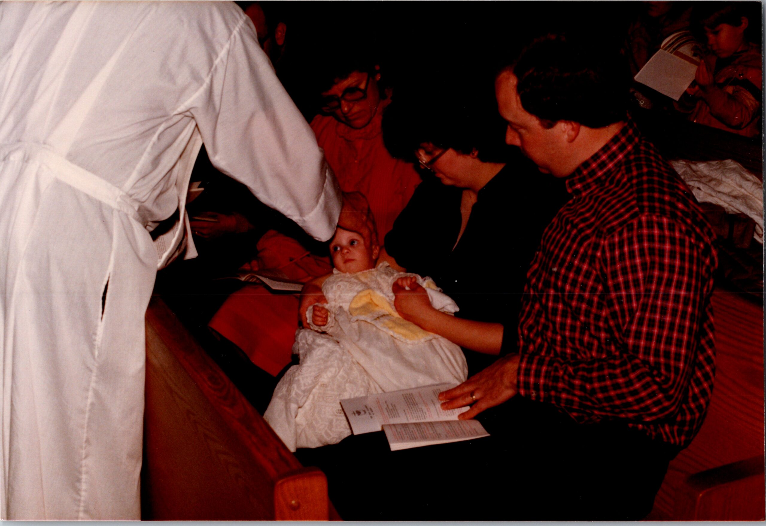 Baby in a white christening gown held by a woman while a man in a red plaid shirt watches, at a baptism ceremony with a priest nearby.