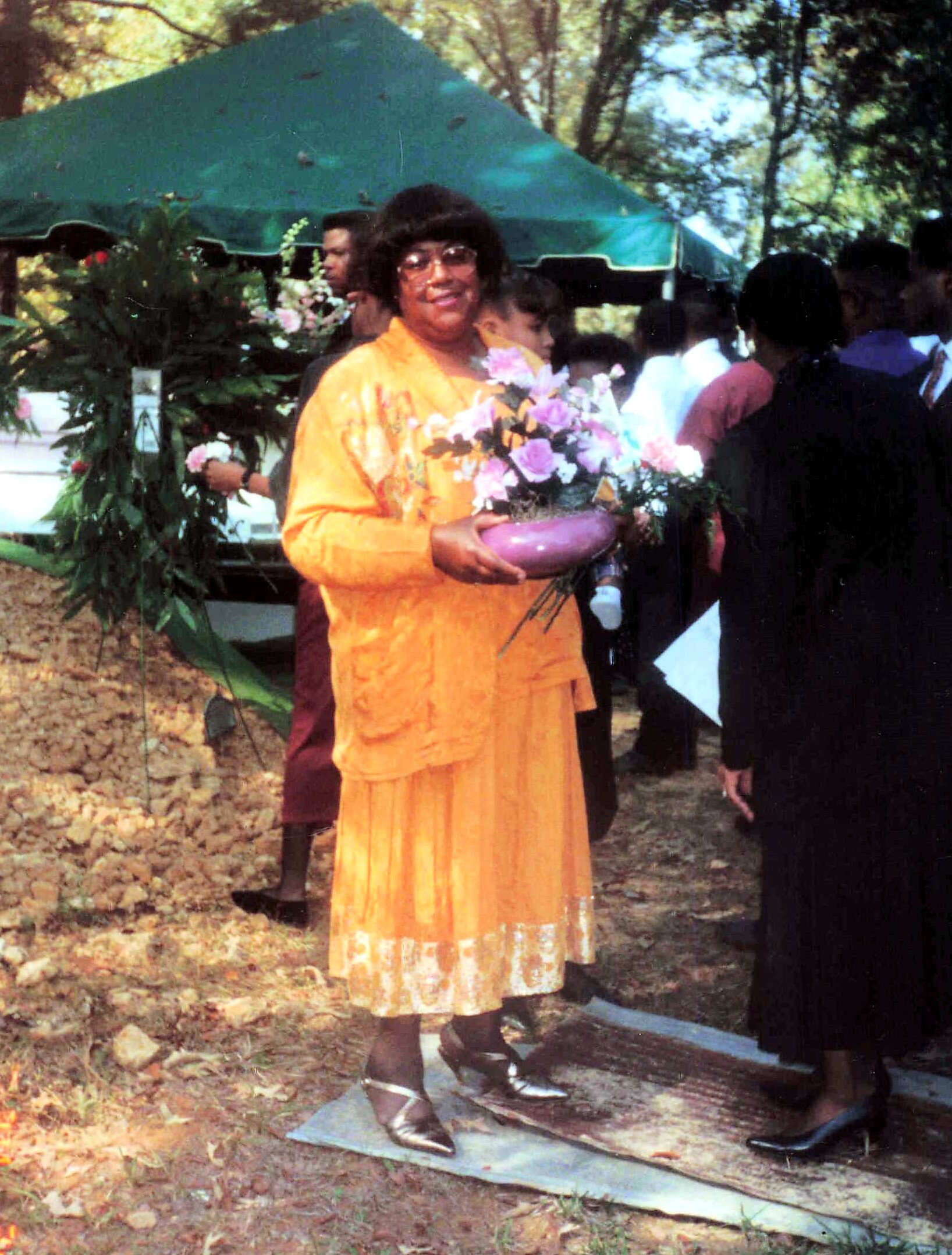 Person in bright orange traditional clothing holding a pink flower bouquet in a purple vase at an outdoor event under a green canopy.