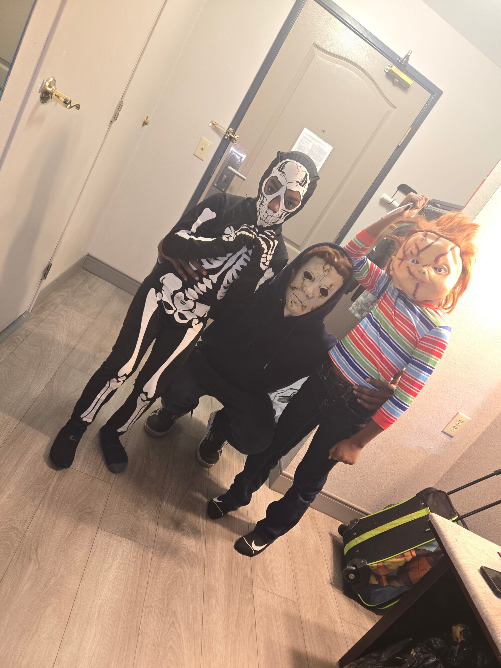Three people in costumes posing in a hallway; one in a skeleton suit, another in a creepy mask, and a third with orange hair and striped shirt taking a selfie in a mirror.