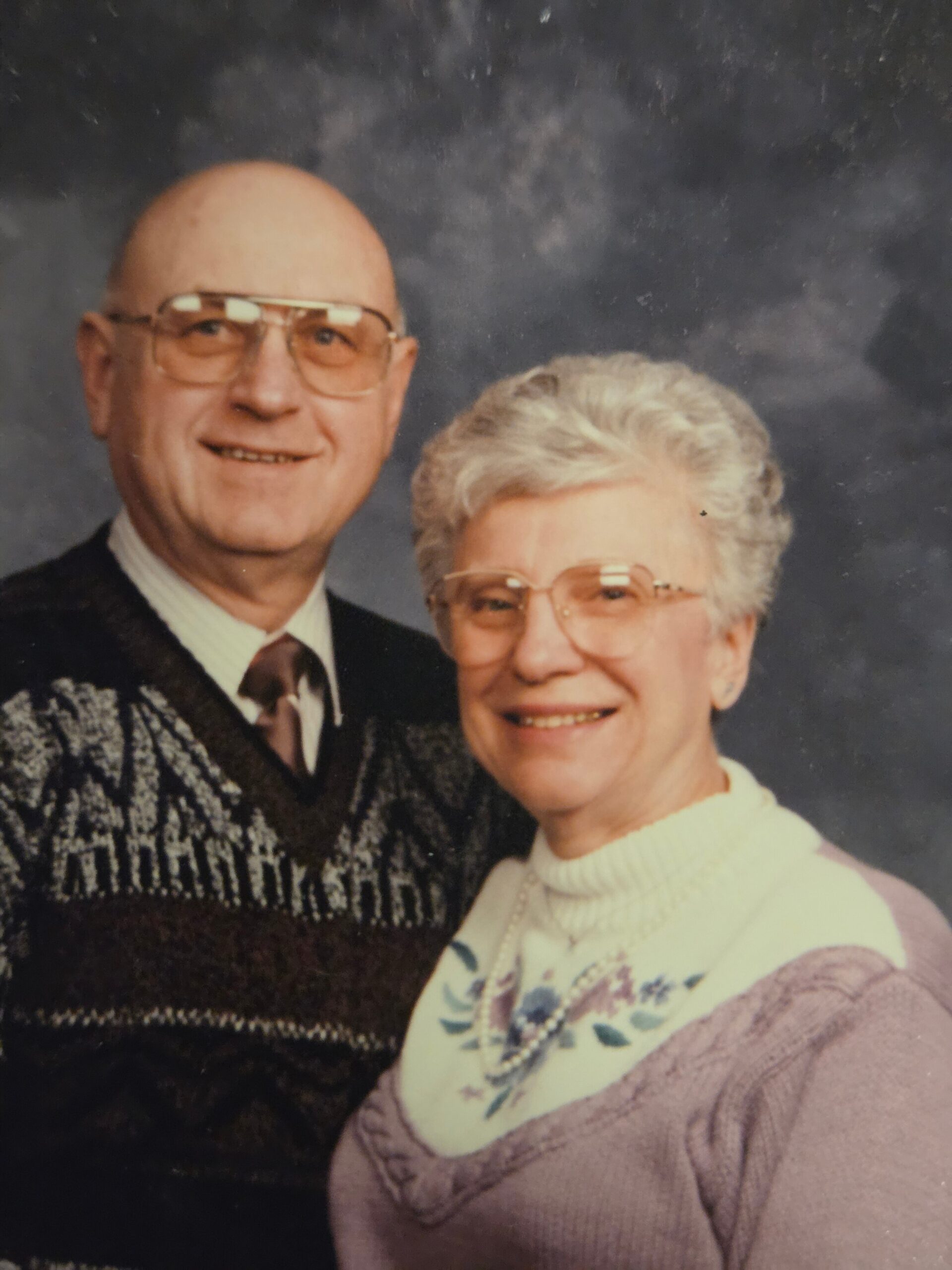 Smiling elderly couple posing for a studio portrait: man with glasses and a patterned sweater, woman with short gray hair and a light sweater.