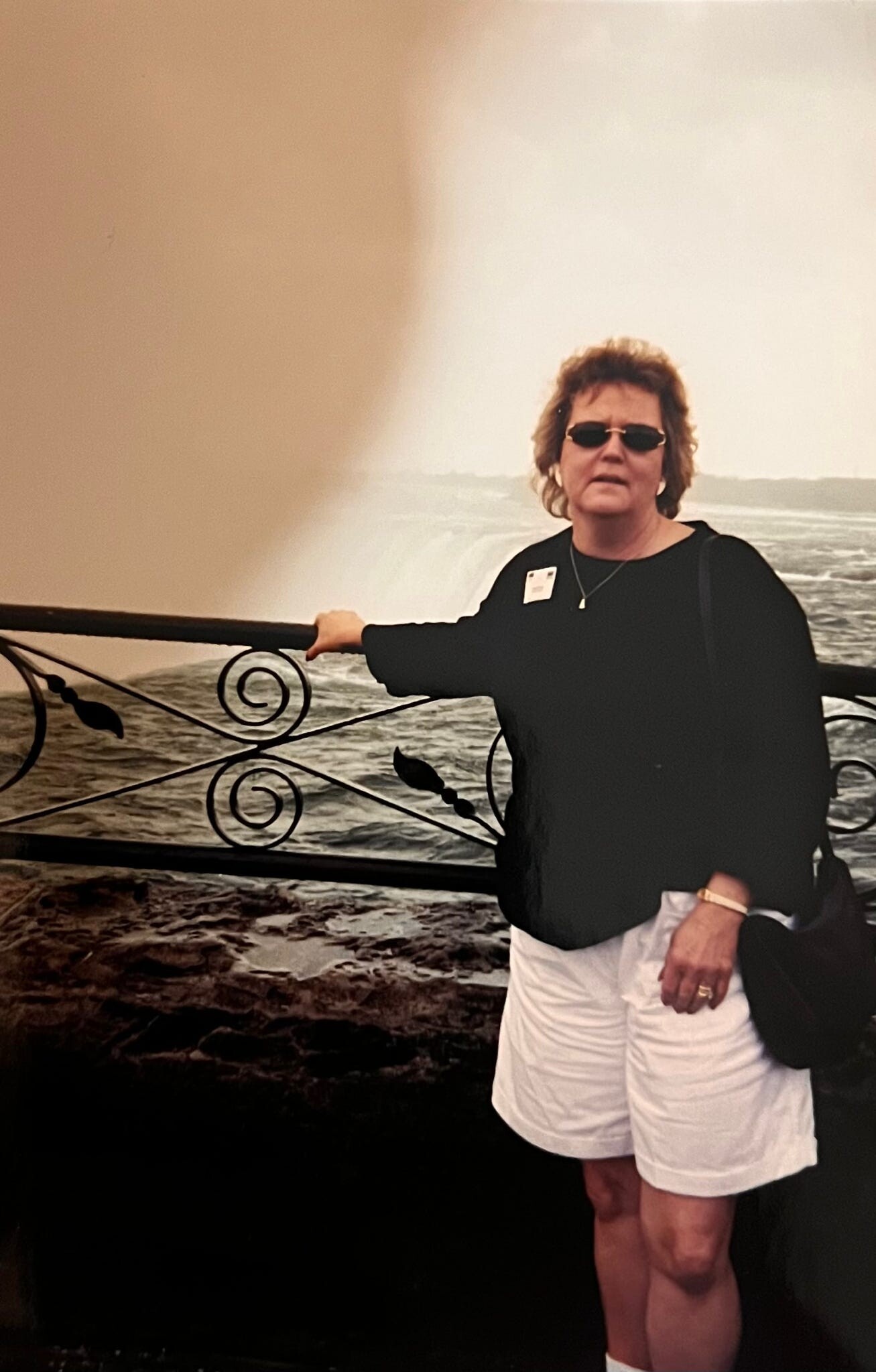 Older woman wearing a black top and white shorts, sunglasses, and a necklace, leaning on a decorative railing by the sea with large waves behind her.