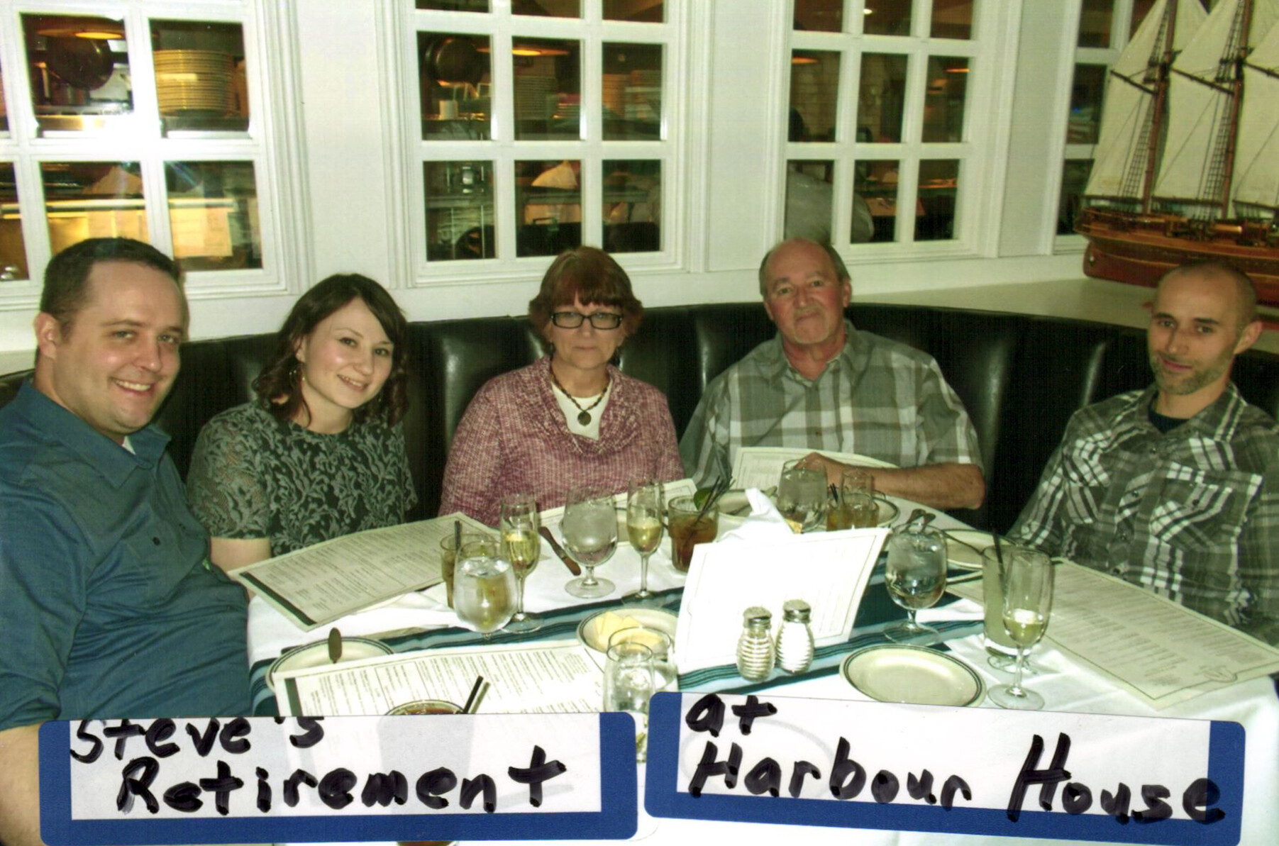 Five adults seated at a restaurant table with drinks and menus; signs read 'Steve's Retirement' and 'Harbour House'.