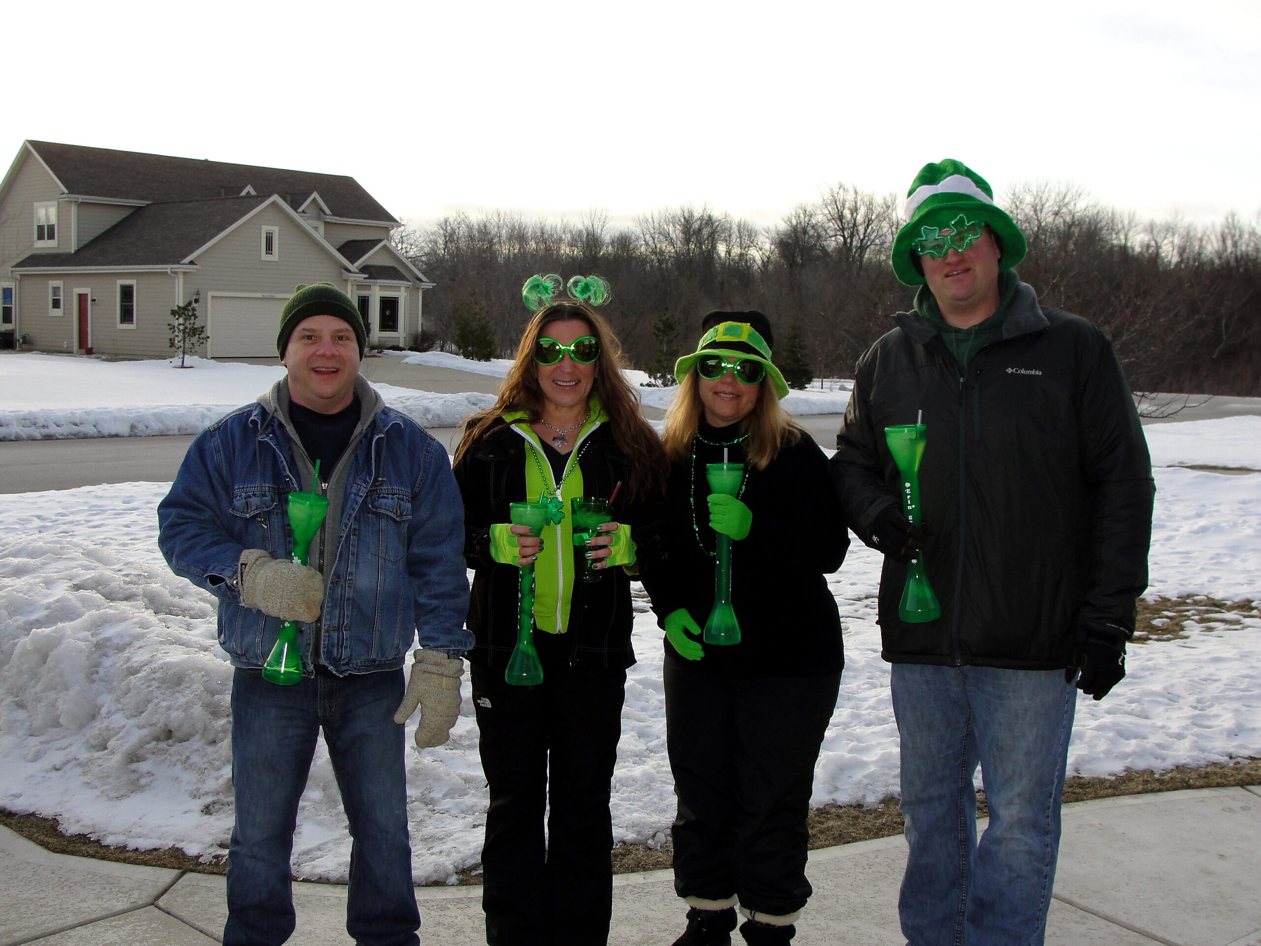 Four adults in winter clothing posing for a group photo outdoors, holding bright green drinks with straws and wearing green accessories for St. Patrick's Day, in a snowy suburban street.