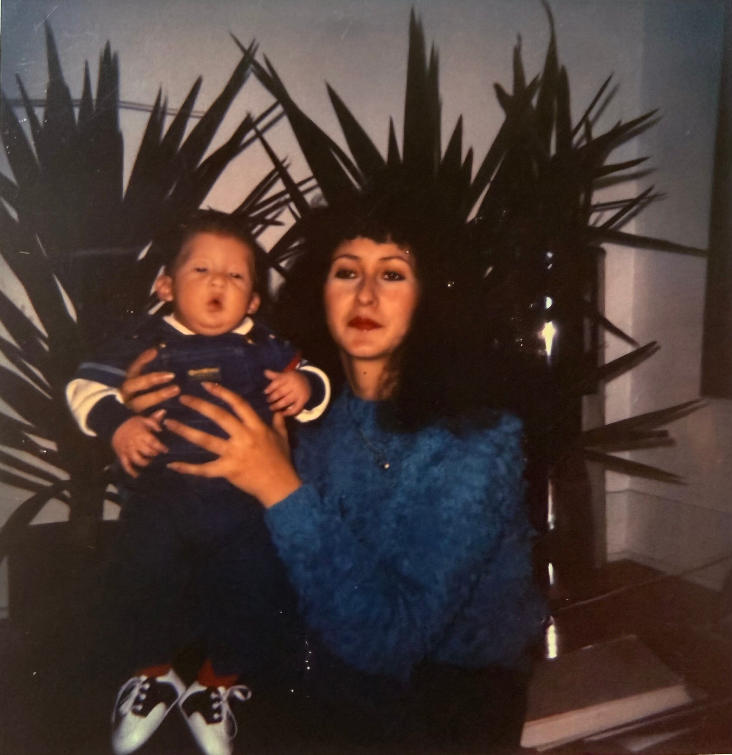A smiling woman in a blue sweater holds a baby in a striped outfit, with a large plant shadow on the wall behind them in a home setting.