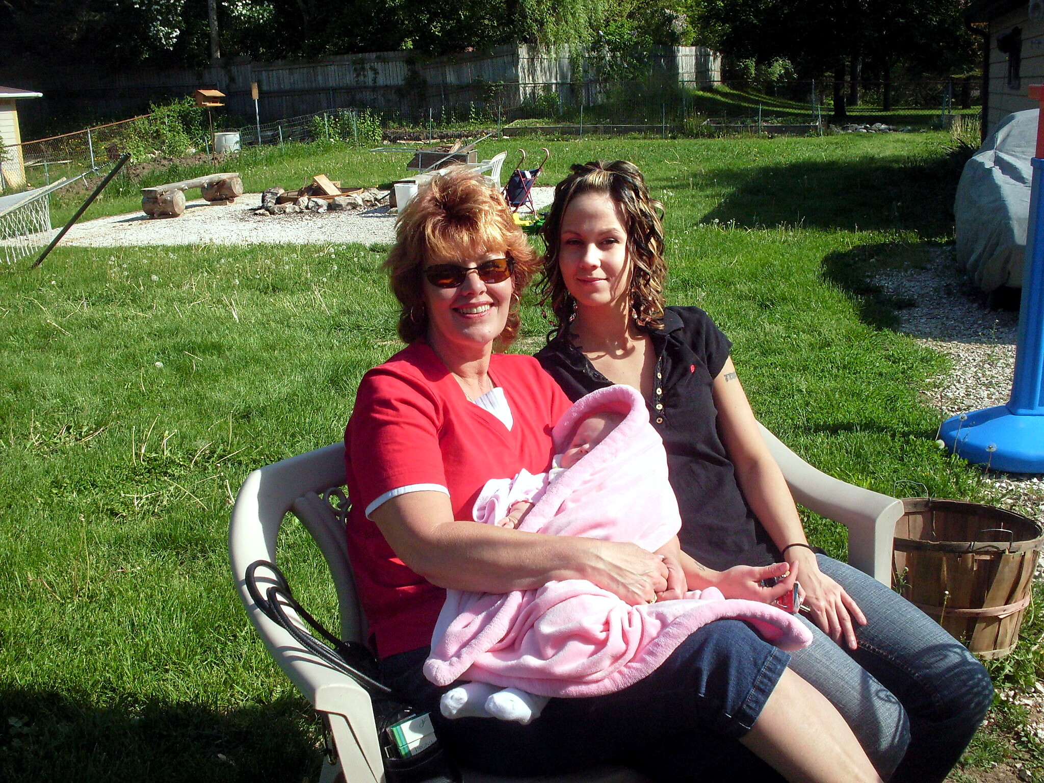 Two smiling women sit in a yard chair outdoors, holding a baby wrapped in a pink blanket. A cluttered backyard with grass and a fire pit is behind them.