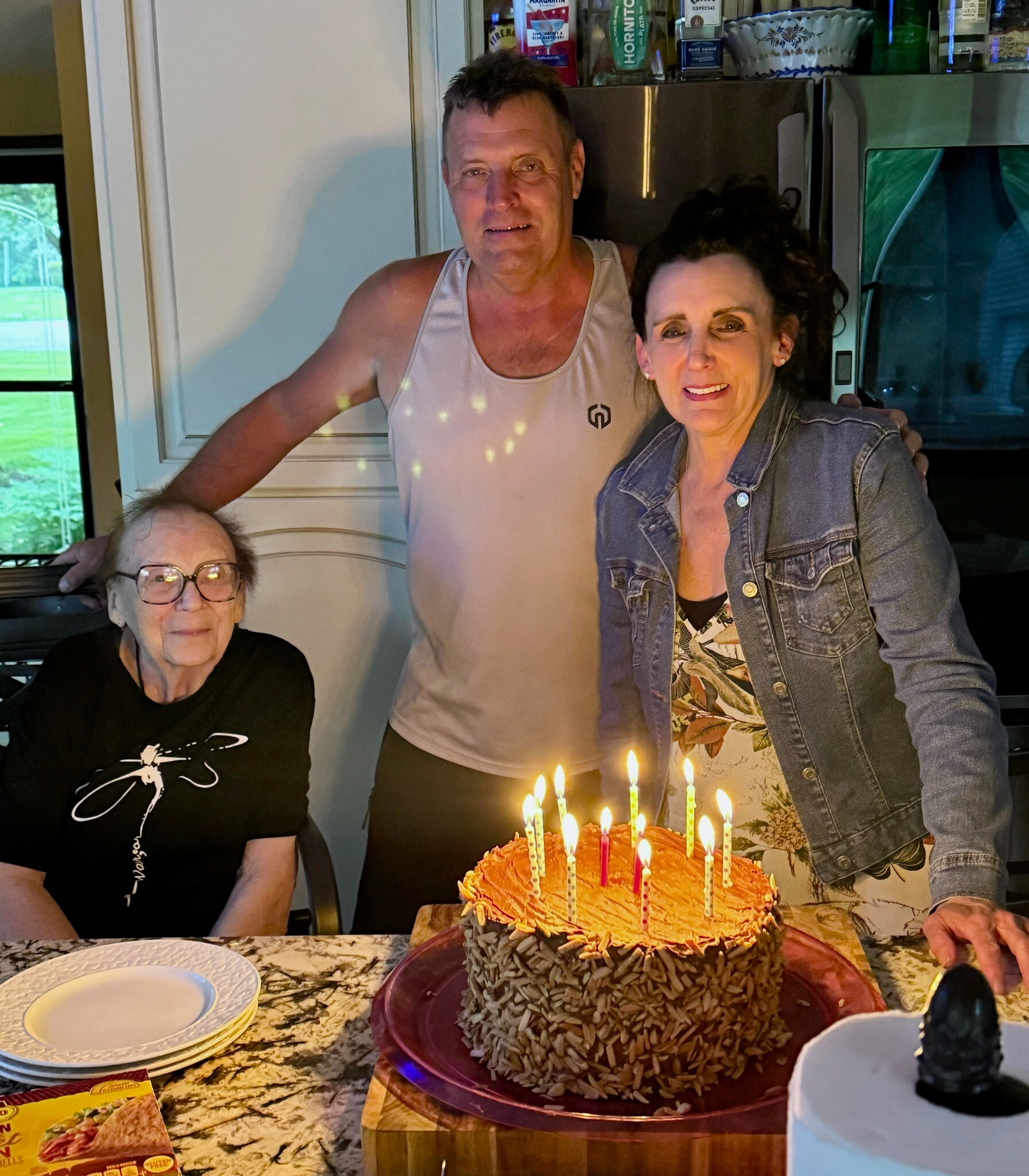 Three adults pose in a kitchen around a birthday cake with multiple lit candles on the counter.