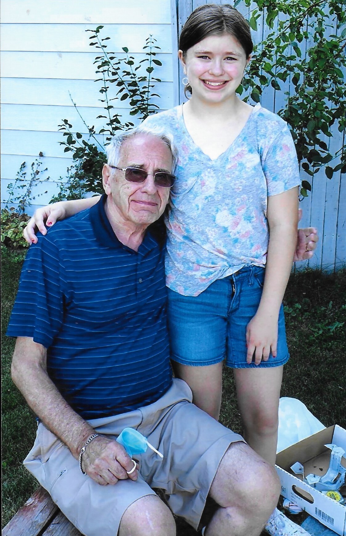 Teenage girl stands beside an elderly man outdoors, arm around his shoulder, both smiling in front of a blue house exterior and plants.