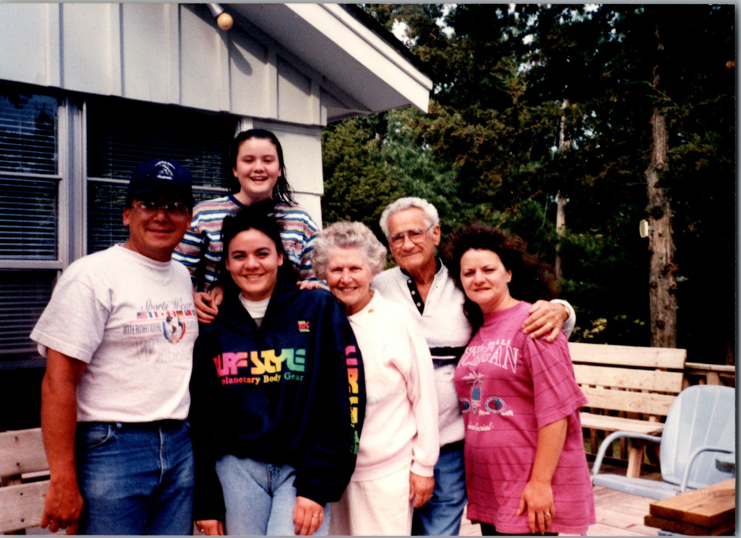 Group of people posing together on a wooden deck outside a house, smiling at the camera (outdoor family photo).