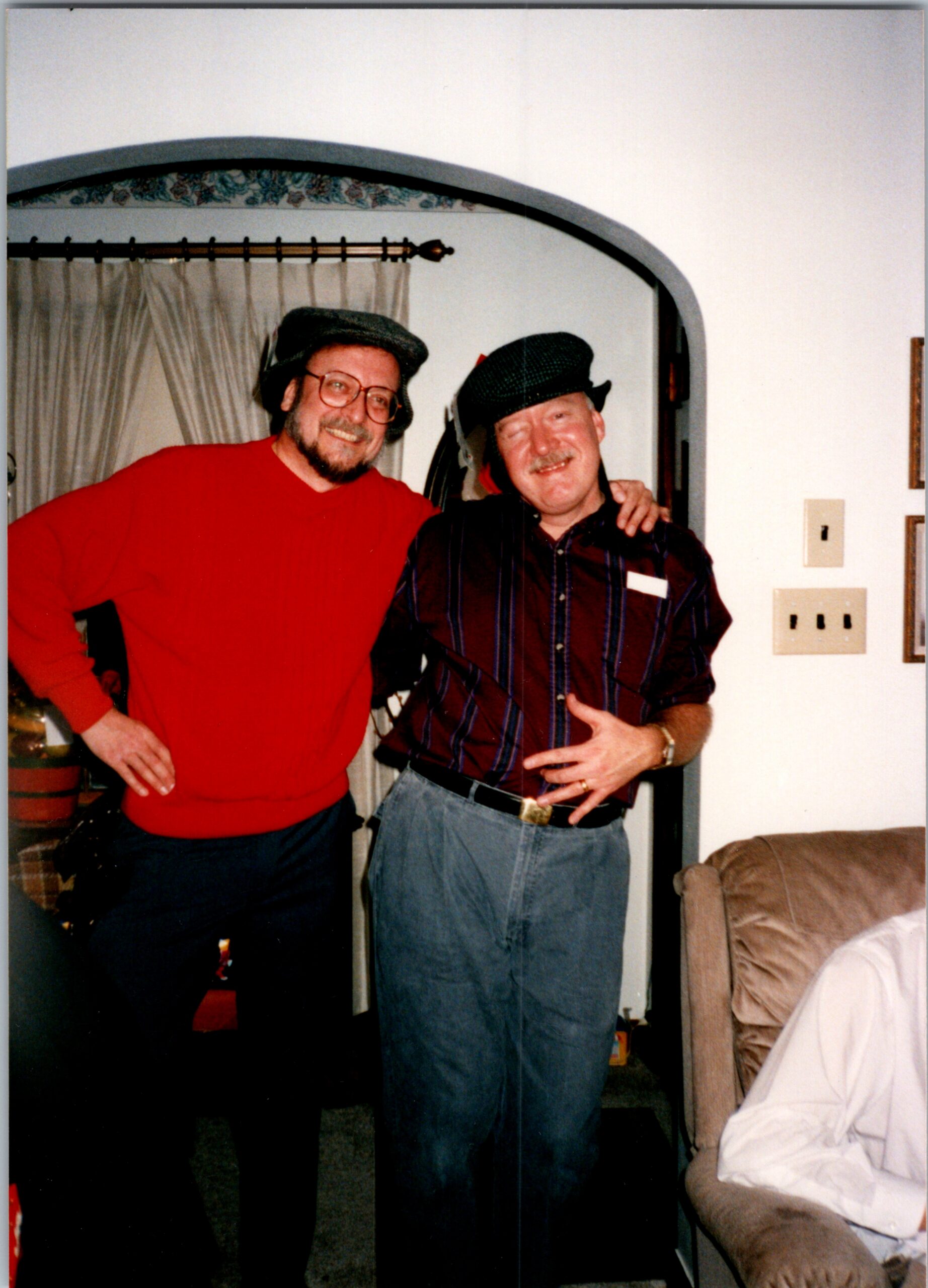 Two men posing together indoors, one in a red sweater and cap and the other in a striped shirt and cap, smiling with arms around each other under a rounded doorway.