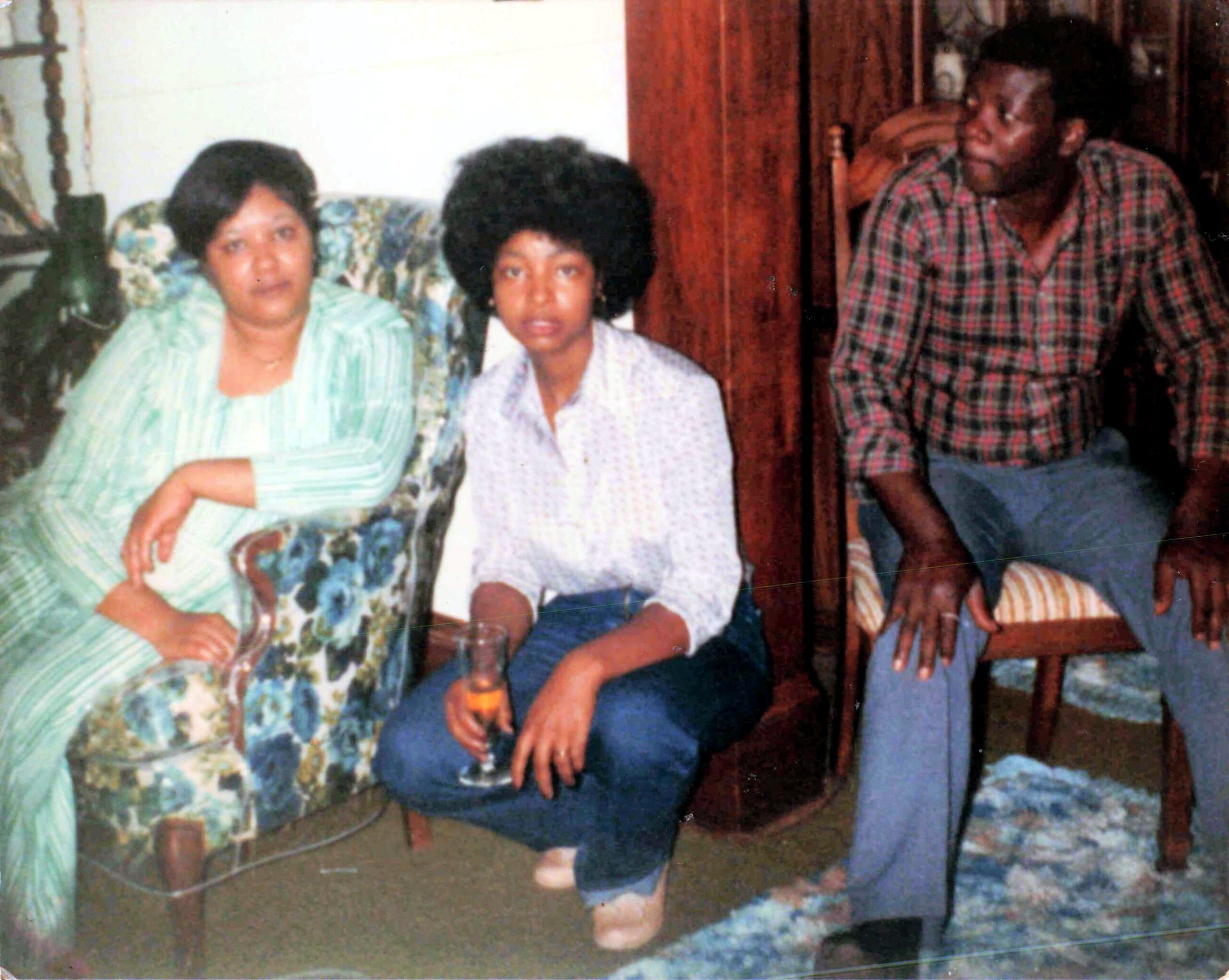 Three people posing for a casual family photo indoors: two women on the left and a man on the right, sitting on and near a chair in a living room.