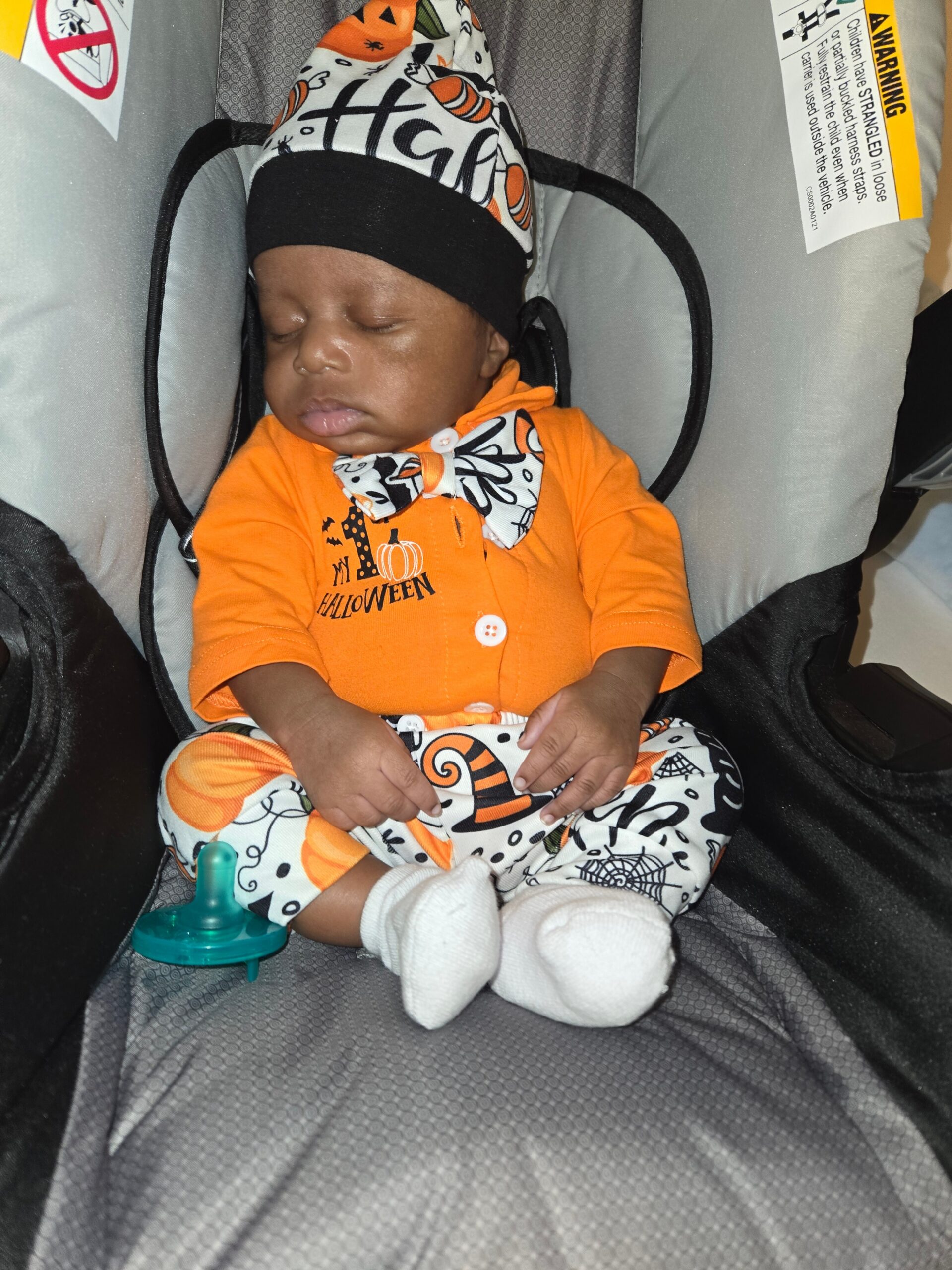 Sleeping infant in an orange Halloween outfit and hat, snug in a gray car seat with a patterned bow tie and socks.