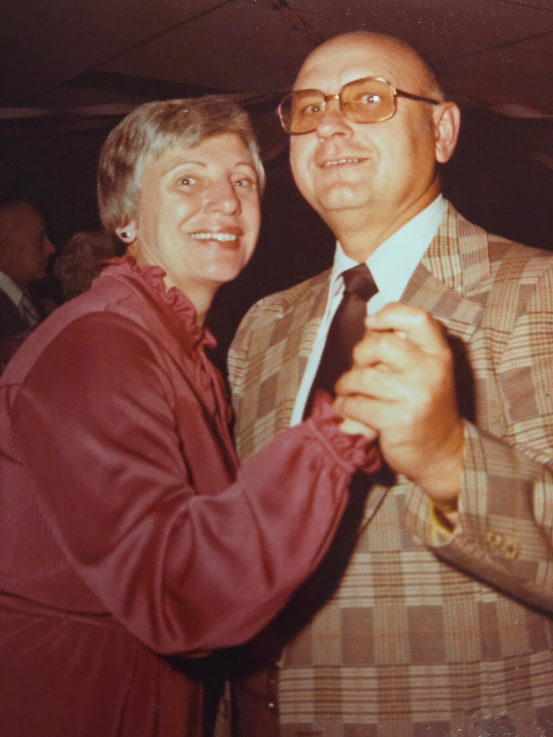 Older couple smiling while dancing at a social event; woman with short gray hair in a burgundy blouse, man in a brown plaid suit and glasses.