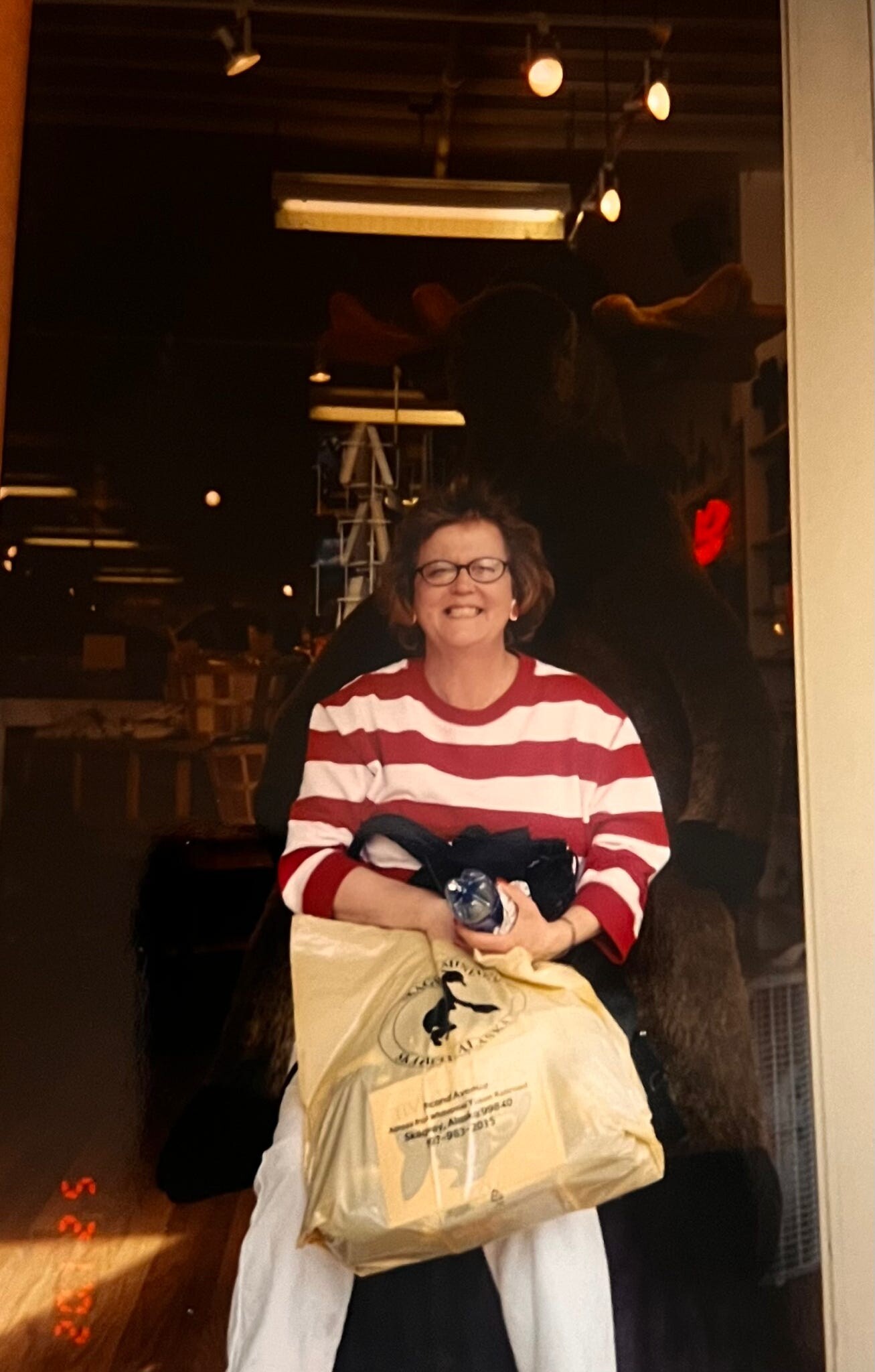 Smiling woman with glasses in a red-and-white striped shirt holding a plastic bag and a water bottle in a shop window display.