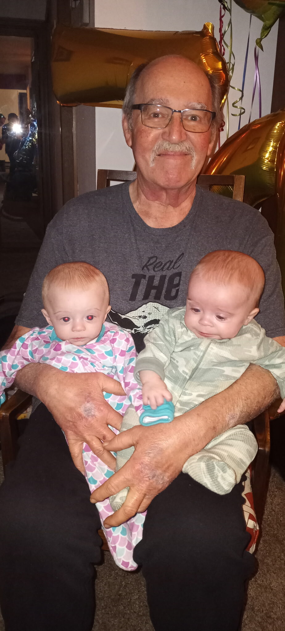 Older man with glasses holding two babies on his lap at a celebration, gold balloons in the background.