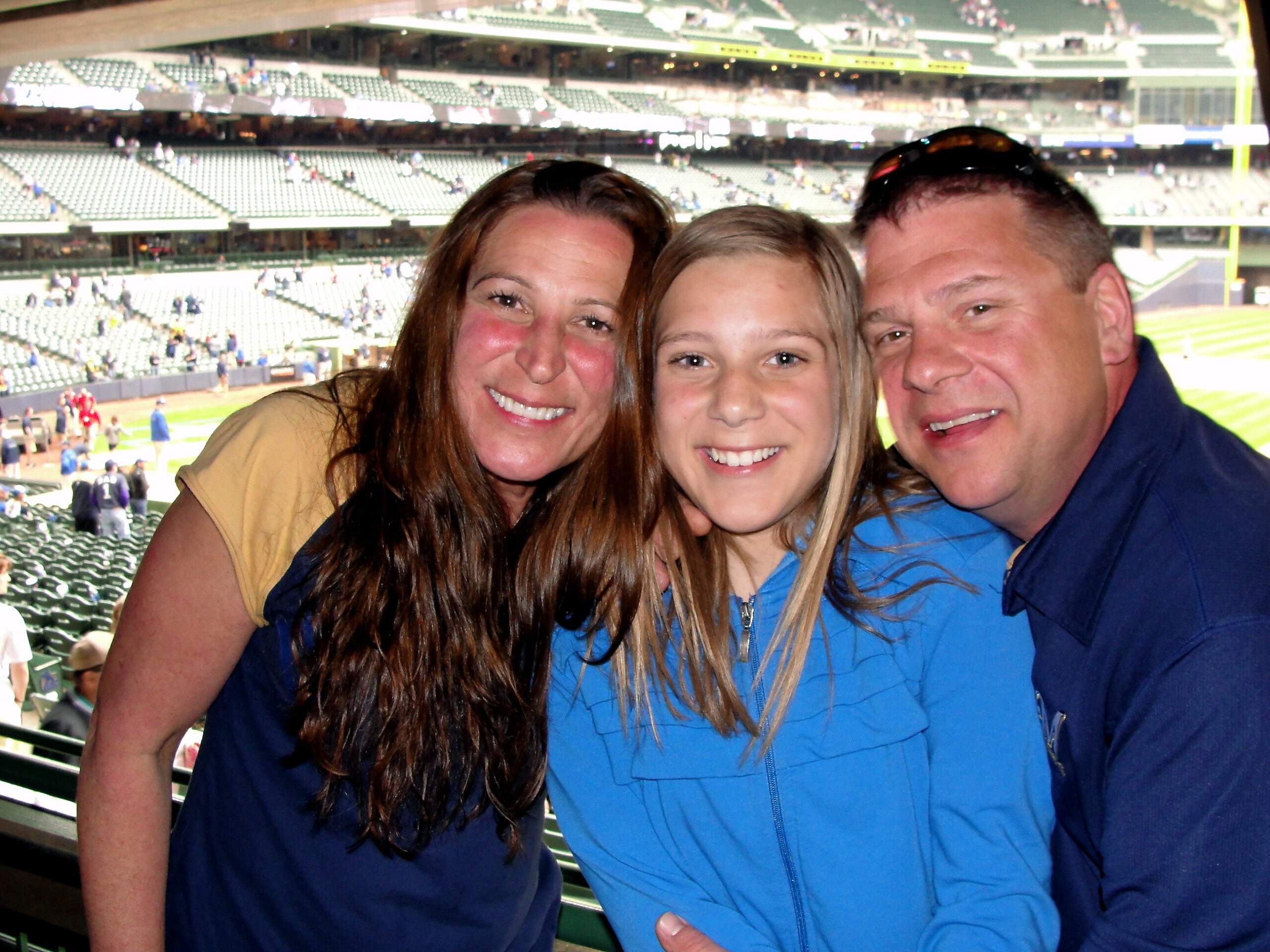 Three smiling people pose for a photo at a baseball stadium, with green seats and the field in the background, wearing jackets.