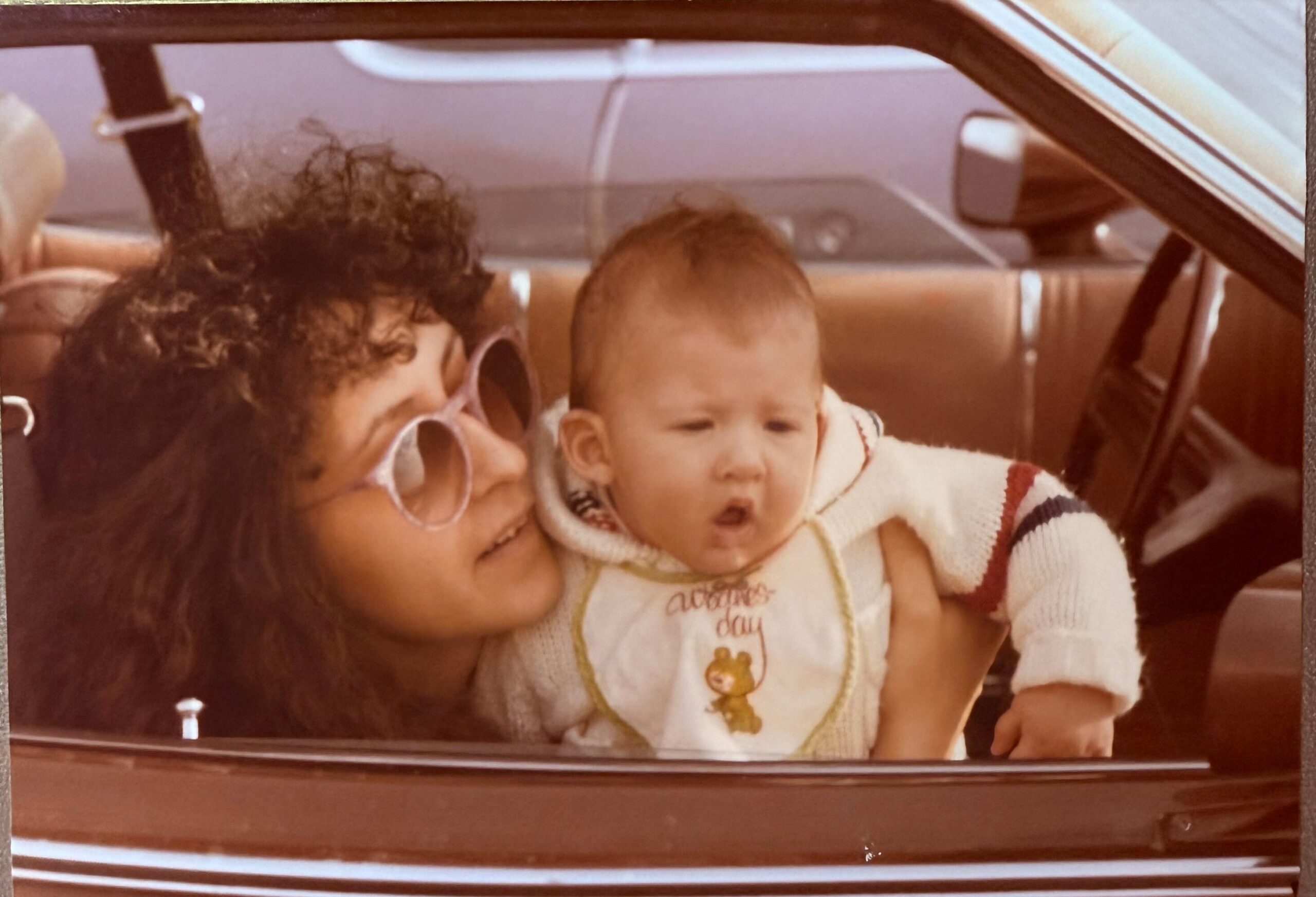 Woman with curly hair and large glasses leaning toward a baby in a striped sweater inside a vintage car interior. The baby looks ahead.