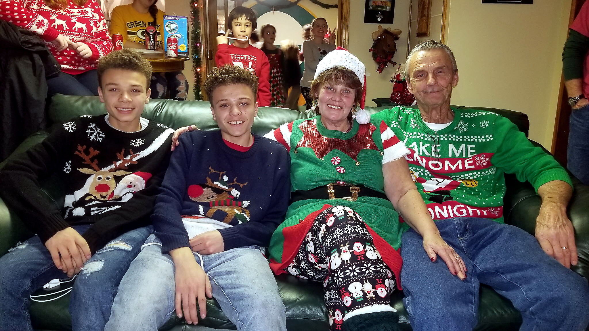 Four people sit on a green couch wearing festive Christmas sweaters, smiling at the camera; background shows holiday decorations and other partygoers.