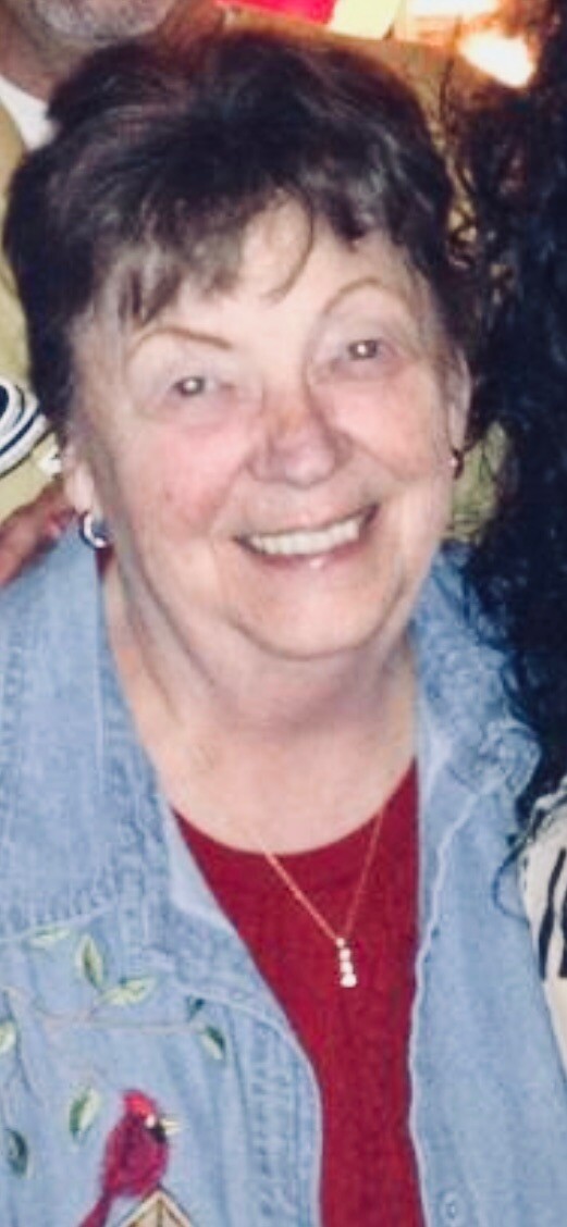 Smiling elderly woman with short dark hair wearing a denim jacket over a red shirt, necklace visible.