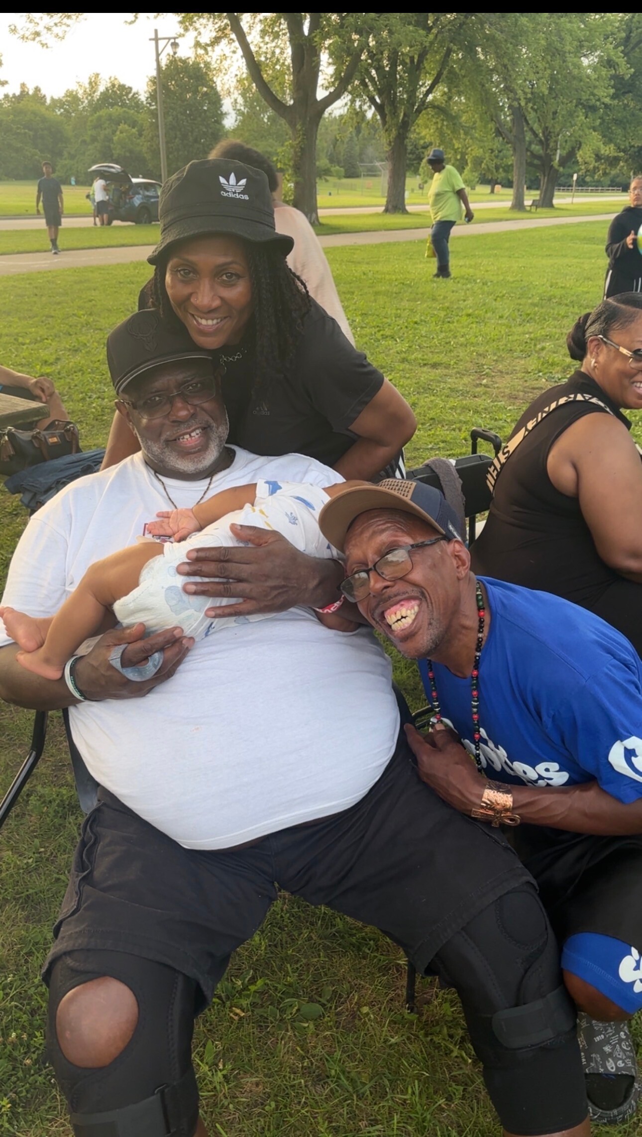 A smiling man in a white shirt holds a baby while a woman hugs him from behind in a park; another man in a blue shirt sits nearby, grinning.